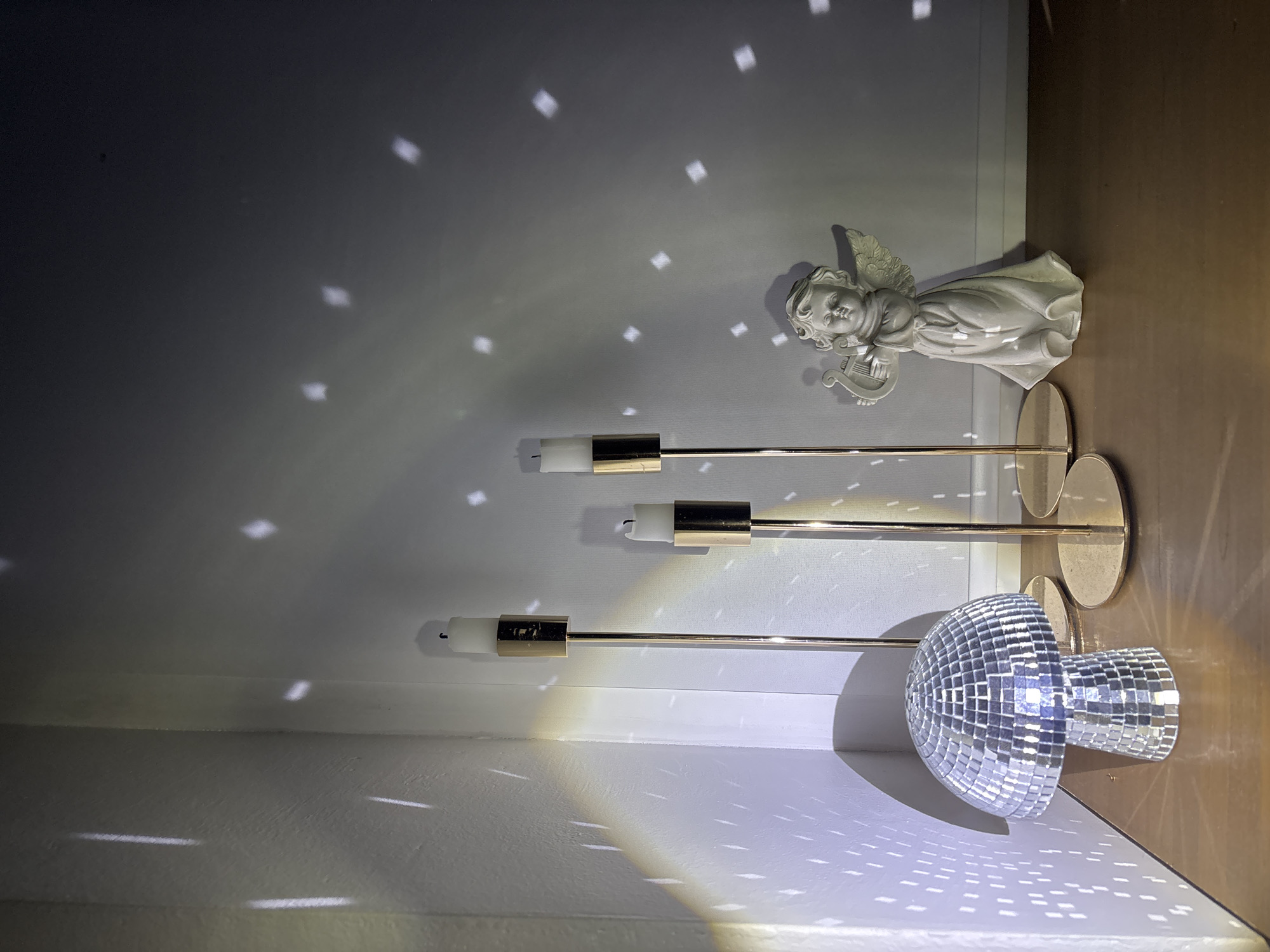 A photograph shows an indoor scene featuring a disco ball, three decorative candleholders, and a small angel figurine. The disco ball is located in the bottom right corner. The candleholders are arranged horizontally, with the tallest one closest to the angel figurine. The angel figurine is positioned near the top right corner. The scene is illuminated by the light reflecting off the disco ball, creating patterns on the walls and ceiling. The location is not identifiable as Chaikovsky, Russia based on the provided image.