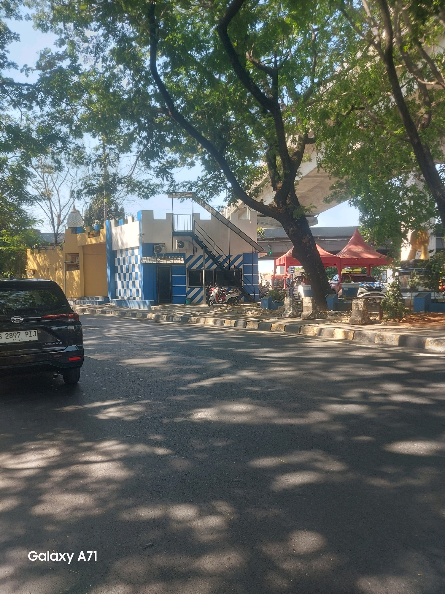 The image shows a street scene in Makassar, Indonesia, on a sunny day. A black Daihatsu Xenia car is parked on the left side of the road, facing away from the camera. The license plate reads "B 2897 PIJ". In the background, there is a building with a light yellow and blue facade, designed with a castle-like parapet. A staircase leads up the side of the building. Red tents are set up near the building, and several motorcycles are parked in front. A pedestrian overpass or bridge is visible in the upper right portion of the image, with trees and foliage obscuring parts of it. The road surface is asphalt, and shadows from trees are cast across it. The overall impression is that of a busy urban environment with elements of architecture and nature.