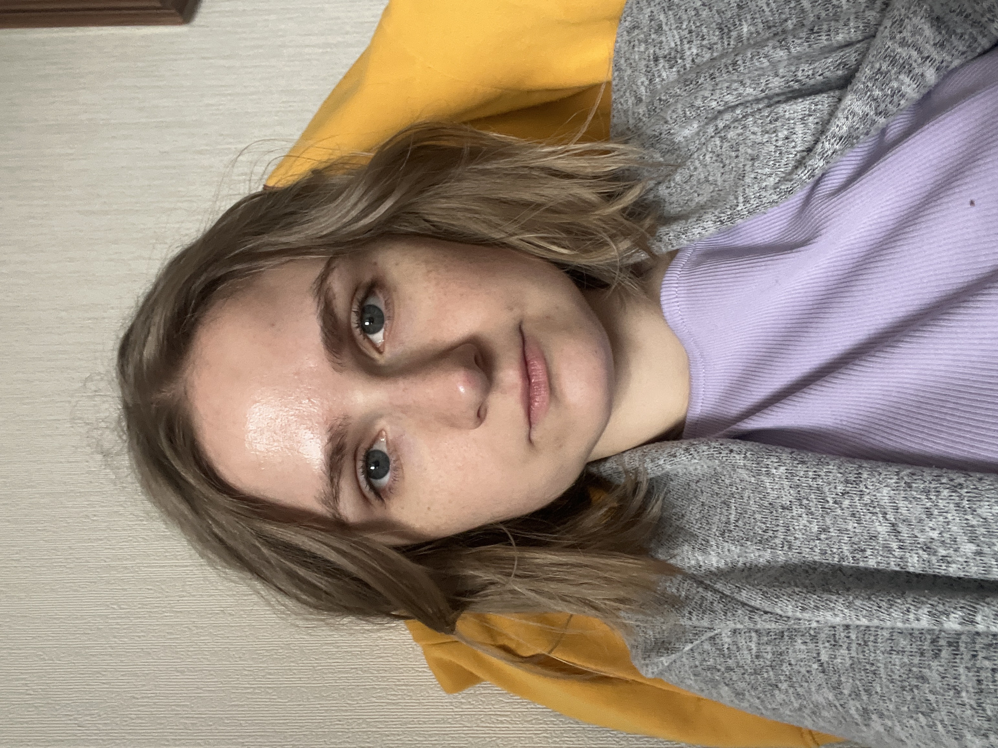 A close-up, slightly angled, indoor photograph depicts a fair-skinned individual with light blonde hair and blue eyes. The person is wearing a light purple ribbed top, covered by a grey knitted cardigan. A yellow garment is partially visible behind the person's head. The background is a light-colored wall with a linear texture. The location, Novosibirsk, Russia, cannot be determined from the image alone.