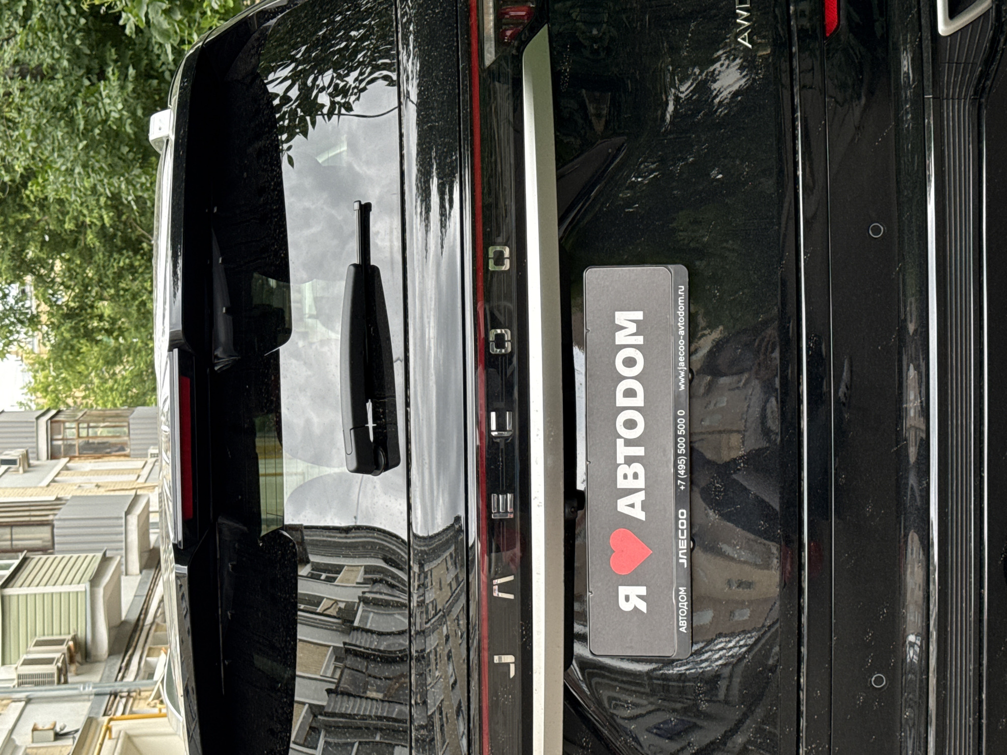 A black SUV is parked on a street in Moscow, Russia. The vehicle has a license plate with Russian text, and the words "Я ♥ АВТОДОМ" are visible on a sticker on the rear of the car, which translates to "I love Avtodom." The car's brand name, "JAEC00," is also visible. The background shows a residential building with balconies and trees. The weather appears to be overcast.
