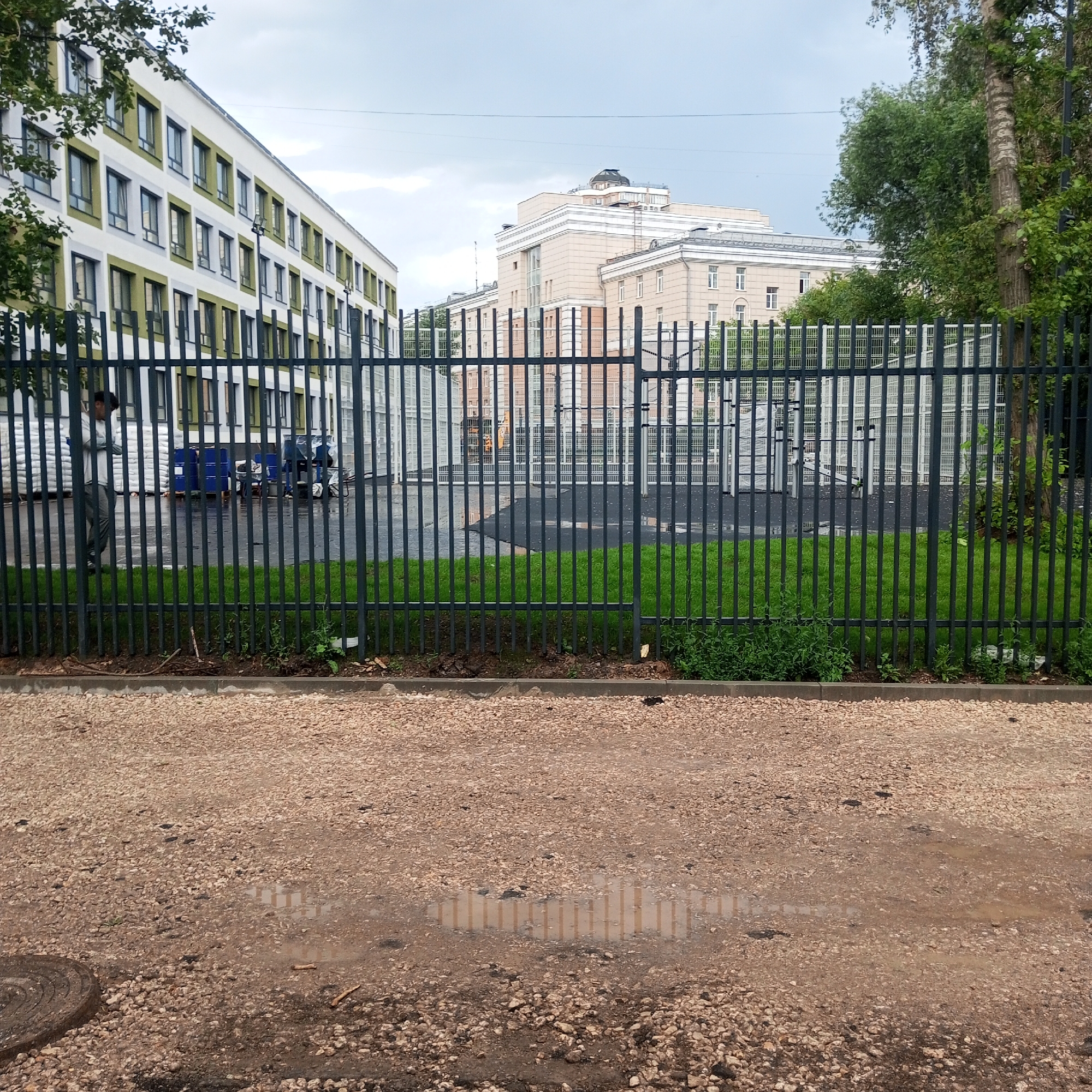 The image shows a view through a metal fence of a schoolyard or educational facility. On the left, a building with white and green facade and many windows is visible. In the center and right, another, larger building with beige and white facade and many windows is seen, possibly also part of the school. Behind the fence, there is a neatly mowed lawn, an asphalt area, and more fencing. On the left side of the yard, a person in a light-colored shirt and dark pants walks away from the viewer. Next to the person are stacked white bags and several blue barrels. The time of day is unclear, but the sky is overcast, suggesting it might be a cloudy or rainy day. The ground in the foreground is covered with gravel and dirt, with puddles reflecting the fence and the sky. A manhole cover is visible in the lower left corner. The setting appears to be urban, given the presence of multiple buildings and infrastructure.