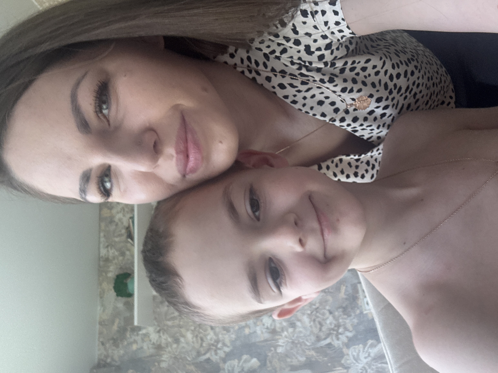 A woman and a young boy are visible in a selfie-style photograph. The woman has light skin, long brown hair, and is wearing a shirt with a black and white cheetah pattern. She is wearing a thin necklace with a pendant. The boy has short brown hair, light skin and is also wearing a thin necklace. The background shows a patterned wallpaper. The location is identified as Раздолье, Russia.