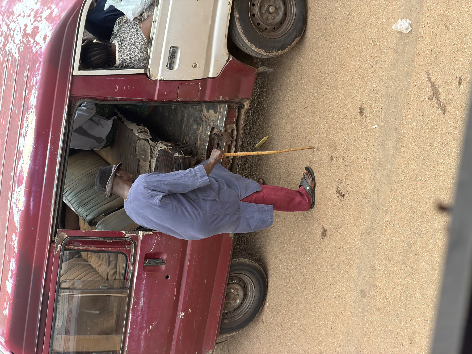 An elderly Black man wearing a dark hat, a blue long-sleeved tunic, and maroon pants, stands on a dirt road next to the open sliding door of a well-worn, maroon and silver minibus in Bukuru, Nigeria. The man holds a wooden walking stick in his right hand and appears to be preparing to enter or exit the vehicle. Inside the van, two other people are visible, one with braided hair wearing a patterned shirt, and another further back in grey clothing. The minibus is in a state of disrepair, with peeling paint, rust around the doors, and worn interior seats. The ground is a sandy, reddish-brown dirt surface with scattered debris, including a small plastic bag and some dark stains. The lighting suggests it is daytime.