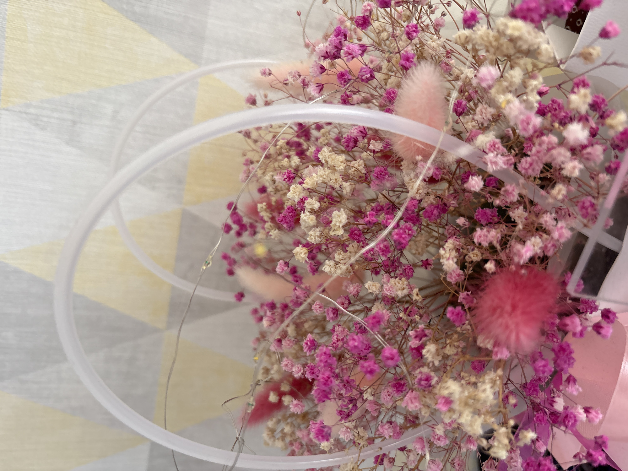 A close-up shot depicts a bouquet of dried flowers in shades of pink and cream, arranged within a transparent, circular frame. The bouquet contains tiny, delicate flowers and fluffy pink seedheads. Thin, barely visible wires are intertwined among the flowers, possibly part of a decorative lighting element. The background consists of a geometric pattern featuring shades of gray, yellow, and white. The context is an indoor setting. The city of Прикамский, Russia, is unrelated to the image content.
