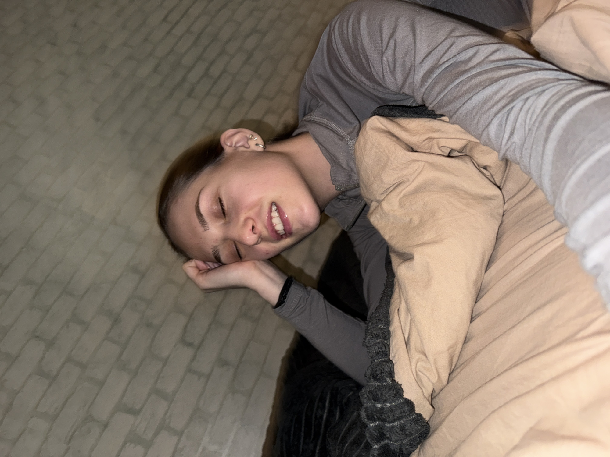 A young woman with fair skin and brown hair is lying on a bed, partially covered by beige and grey blankets. She is wearing a grey long-sleeved shirt. Her head rests on her left hand, and she appears to be smiling with her eyes closed. The background suggests a neutral-toned wall or surface. The location is unknown and cannot be determined from the image. The image does not provide any information about Sofia, Bulgaria.