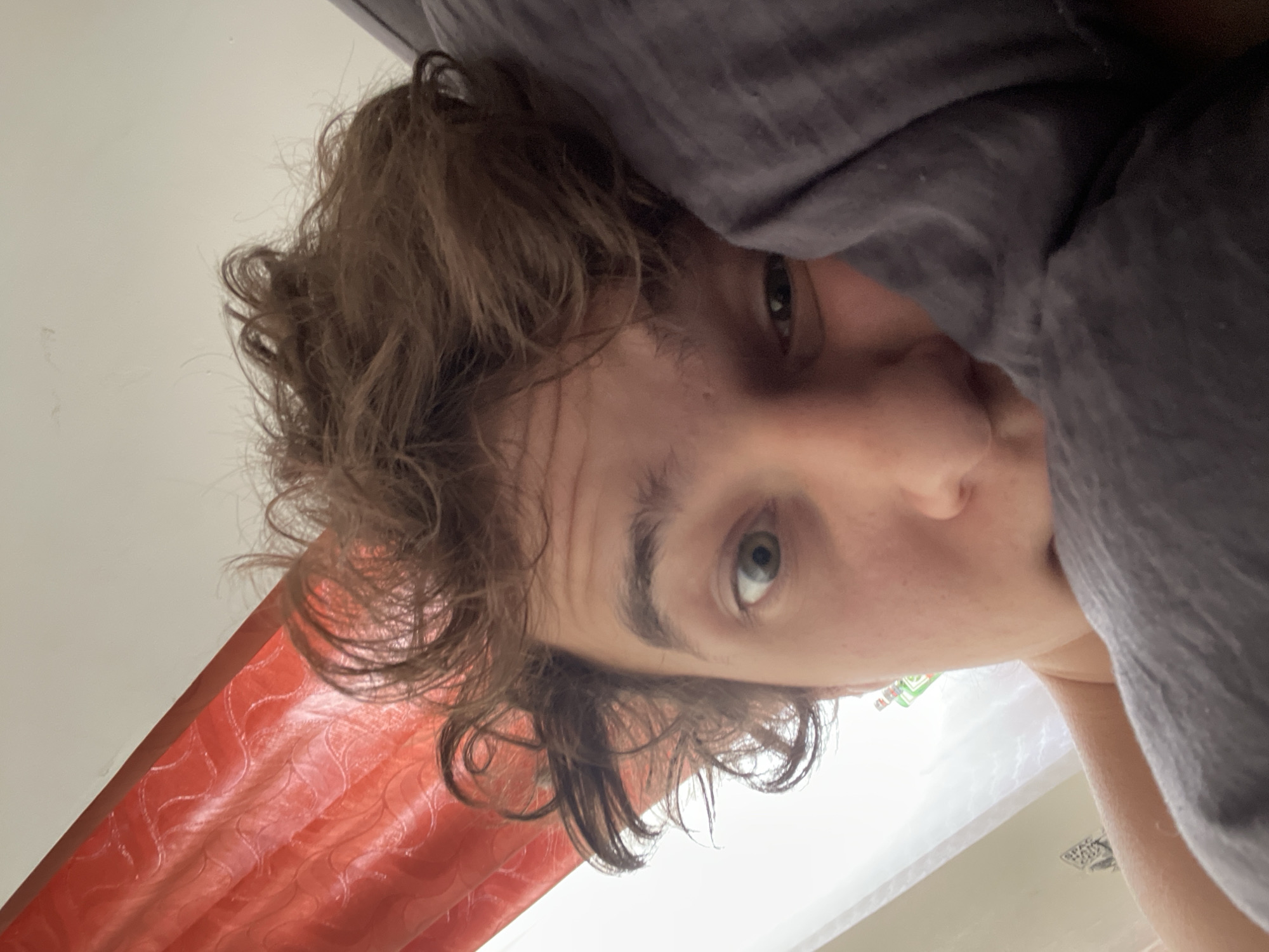 A young adult with light brown, curly hair is partially obscured by a gray fabric, possibly a blanket. The person has light skin and a single visible eye which is green. An orange curtain is visible in the lower left corner of the image. The location is not identifiable as Krasnoyarsk, Russia from the provided image.