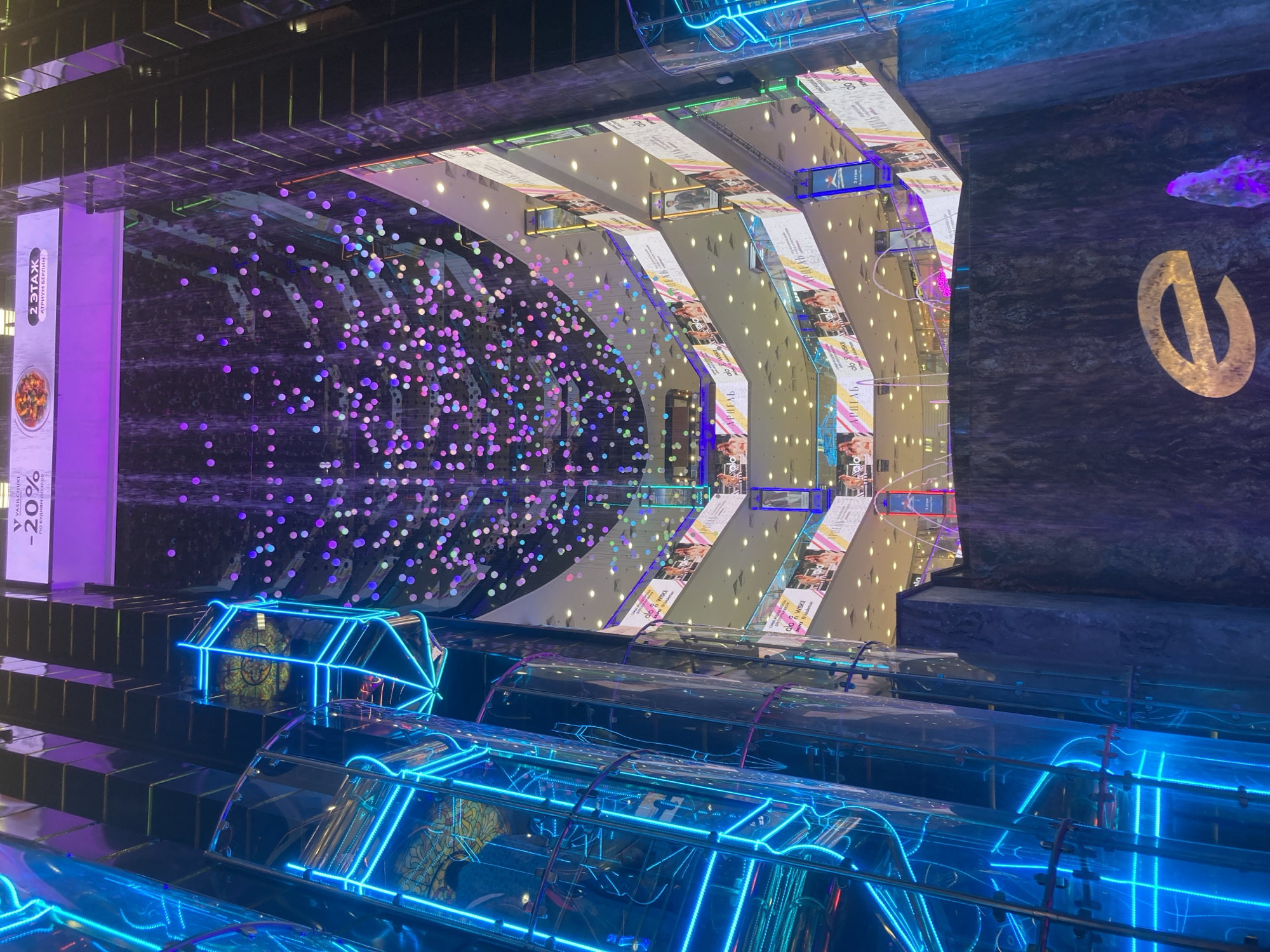 The image depicts the interior of the Atrium Shopping Mall in Moscow, Russia, specifically within the "Atrium Berlin" section, as indicated by a prominent sign reading "2 ЭТАЖ АТРИУМ БЕРЛИН." The scene is a vertical, multi-story view of a brightly lit commercial atrium, characterized by modern architecture and vibrant artificial lighting.
On the left, a stack of at least three glass-encased elevators ascends vertically, each outlined with bright blue neon light strips, creating a futuristic aesthetic. Reflections are visible on their clear surfaces. To the right, a darker, multi-tiered structure, likely housing escalators, features reflective, possibly metallic, surfaces.
The central void of the atrium is adorned from above by a dense canopy of numerous small, suspended spherical lights, glowing in varied hues of purple, pink, blue, and green, creating a shimmering, rain-like effect. Below this light canopy, a large indoor waterfall cascades down a dark, textured wall. At the base of this wall, a prominent golden 'e' logo is illuminated. In the immediate foreground, a water feature, possibly a fountain, emits water illuminated with purple light.
Multiple shopping levels encircle the atrium, each defined by white barriers or railings. Along these levels, numerous large white banners display advertisements, predominantly featuring female models and product imagery. Text on these banners is in Cyrillic, with visible terms such as "АРДЕЛЬ" and "Wisca." One banner near the top left advertises a restaurant with "V VASILCHUKI -20% по будням до 17:00" (Vasilchuki -20% on weekdays until 17:00). Indistinct human figures are sparsely visible on the upper levels, appearing as small silhouettes against the interior lighting. The overall environment is clean, well-maintained, and designed for retail, utilizing glass, reflective materials, and dynamic lighting to create an expansive and energetic atmosphere.