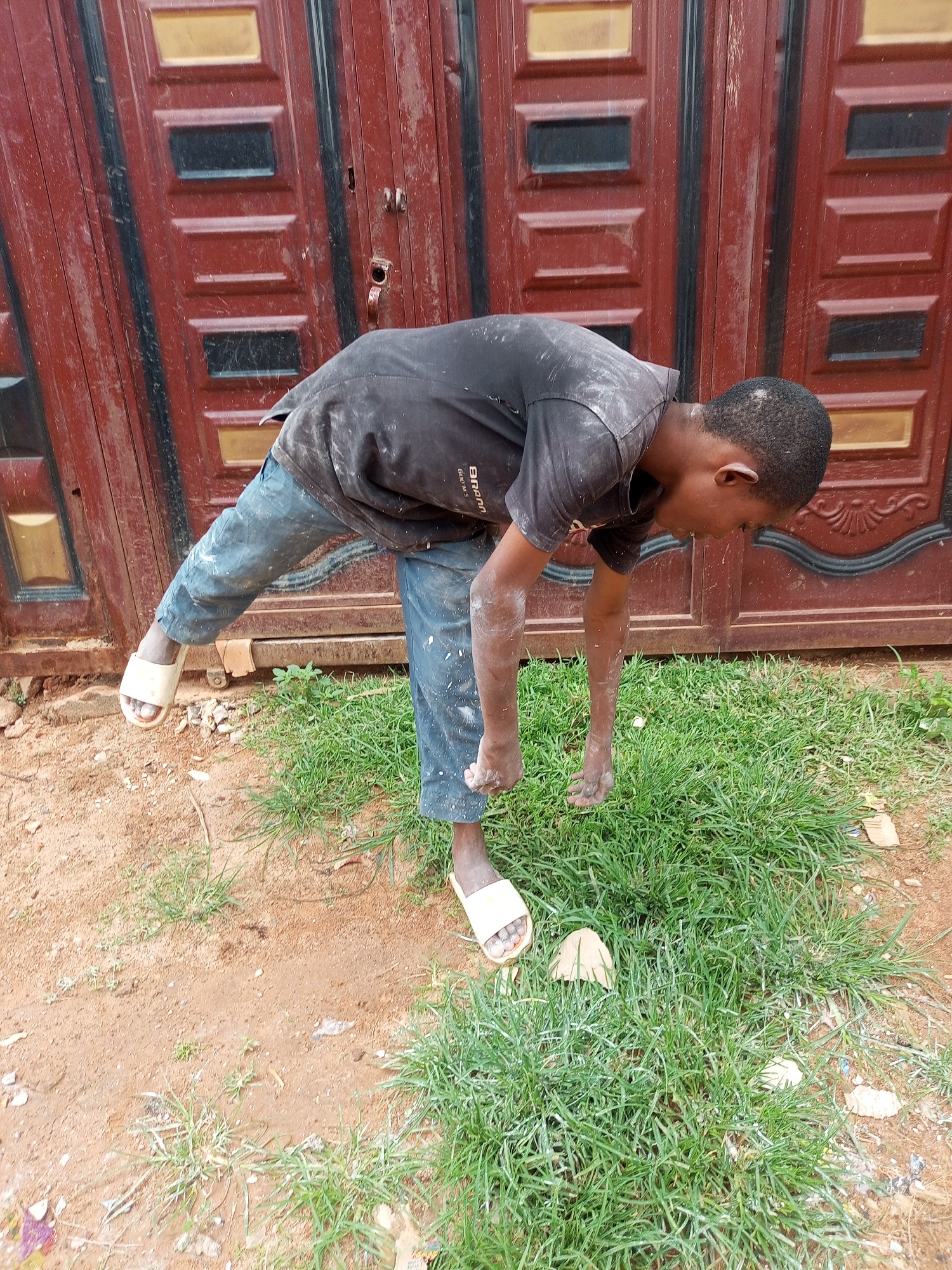 A young boy, appearing to be around 10-12 years old, stands bent over in front of a dark red metal door with rectangular panels. He is wearing a dark t-shirt covered in white powder and light blue pants, also speckled with white powder. He has on light-colored sandals. His left leg is extended backward, and his right leg is bent at the knee. His hands are close to the ground, reaching into a patch of green grass. The ground is a mixture of brown dirt and grass. The overall scene suggests an outdoor setting, possibly in a residential area. The presence of white powder on his clothing and skin indicates he may have been involved in an activity like painting or construction. The backdrop of the door and the surrounding environment are consistent with an urban or suburban setting in Nigeria.