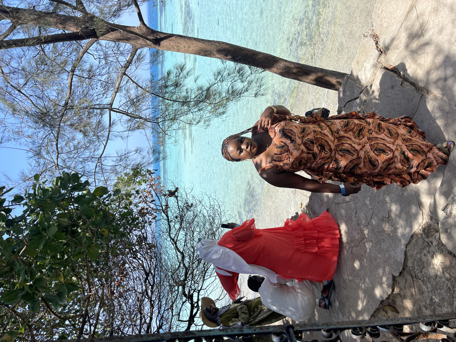 The image captures an outdoor coastal scene in Bububu, Tanzania, under a clear blue sky. Four individuals are present on an uneven concrete surface adjacent to clear, turquoise ocean water. In the foreground, a woman with braided hair, wearing a sleeveless, floor-length dress patterned in orange, brown, and black, stands looking directly at the viewer. She has a blue wristband on her left wrist and a dark bag slung across her body. To her left, a person in a bright red, ruffled dress and white hijab faces the water, holding a black smartphone horizontally, actively capturing imagery. Further left, a person in an olive-green garment and a light-colored wide-brimmed hat over a hijab is partially visible. Between these two, a fourth individual, dressed in white, crouches on the concrete surface, also facing the water. Tree branches with green foliage and bare branches frame the scene from the left and right, casting shadows on the ground.
