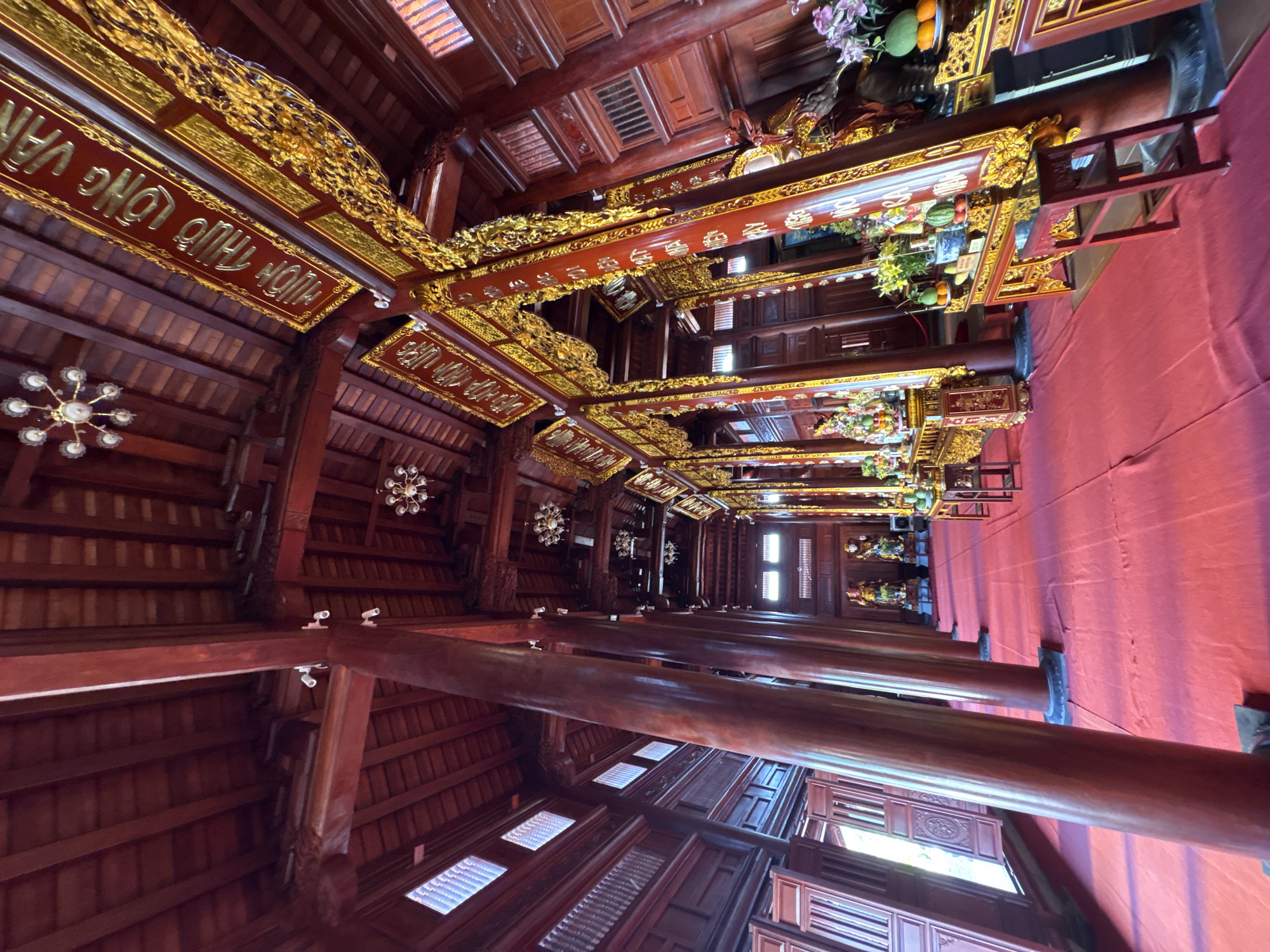 The image captures the interior of a traditional Vietnamese structure, possibly a temple or communal house, located in Nam Nha Trang Ward, Vietnam. The building showcases intricate wooden architecture, with dark red-stained beams and pillars supporting the roof. Golden decorative elements are prominent, adorning the beams and roof structures. The floor is covered with a red carpet or cloth. Several objects, including statues and offerings, can be seen on elevated platforms or altars. The lighting is a combination of natural light filtering through the windows and artificial lighting from chandeliers. The building has a long, narrow structure with pillars running down the length of the room.