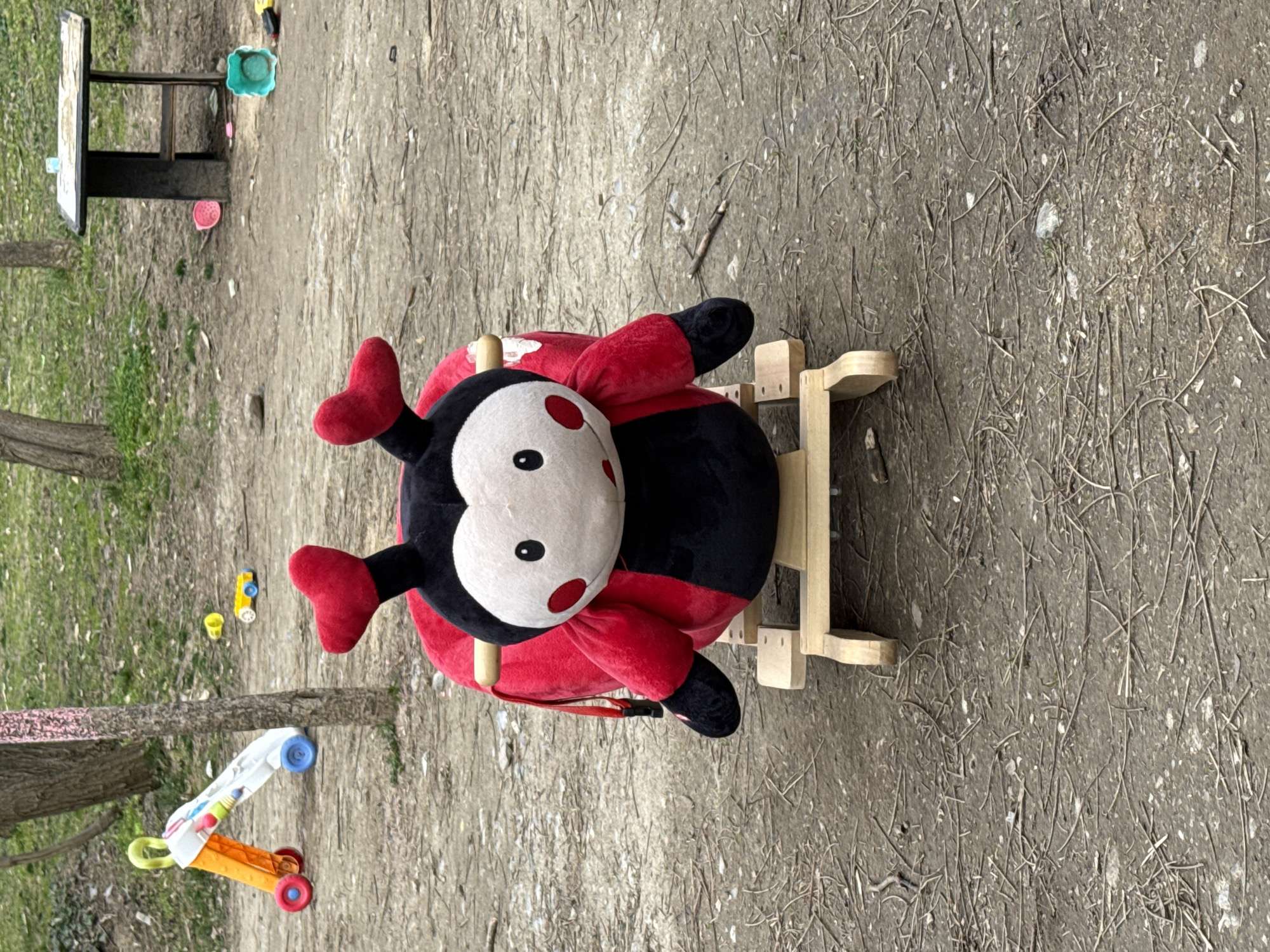 The scene depicts an outdoor play area in a park within Sofia, Bulgaria. A large, plush ladybug rocker, primarily red and black with a white face, black eyes, and red cheek spots, stands centrally on a light-colored wooden rocker base. The ladybug features red heart-shaped antennae and a visible wooden handle on its back. The ground is a mixture of compacted dirt and soil, extensively covered with numerous dried twigs, small branches, and scattered small stones.
In the midground to the left, a multi-colored baby walker, predominantly white and orange with red and blue wheels, is present near a tree trunk. Further left, a small yellow plastic cup and a small yellow toy car are resting on the ground. A section of the tree trunk displays faint pink coloration.
To the right in the midground, a small, dark-stained wooden table with a lighter-colored top is visible. Surrounding the table are several brightly colored plastic children's toys, including a pink plastic bucket, a teal plastic bucket, and various other small plastic items. The background consists of sparse, dull green grass patches and additional tree trunks with bare branches. No individuals are present in the scene.