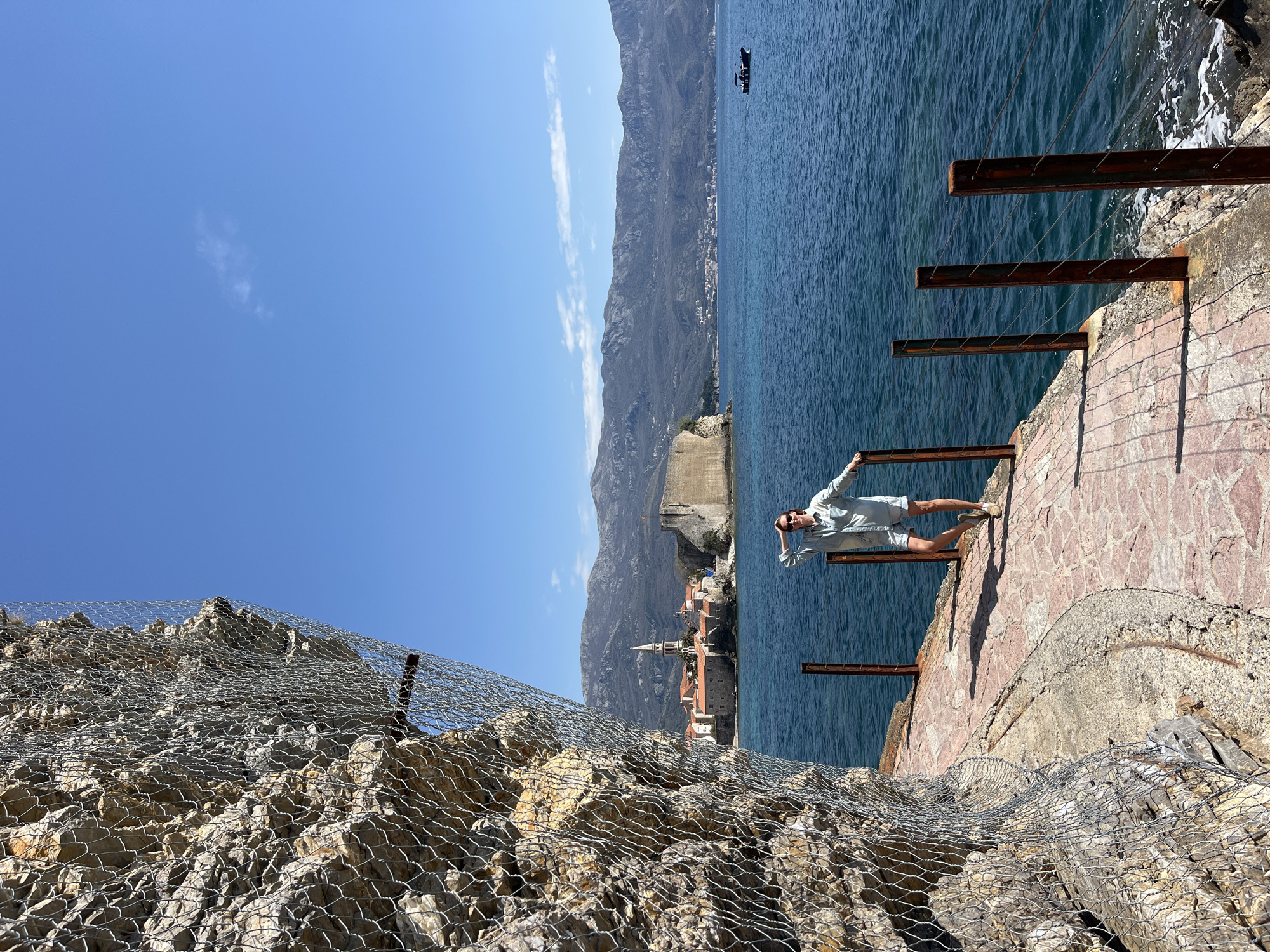 The image shows a person standing on a stone pier with metal supports overlooking the Adriatic Sea. Behind the person are the buildings of Sveti Stefan, a small islet and 5-star hotel resort near Budva, Montenegro, with a mountainous background. To the left of the pier, a steep rock face is covered with wire mesh. The sky is blue with a few clouds.