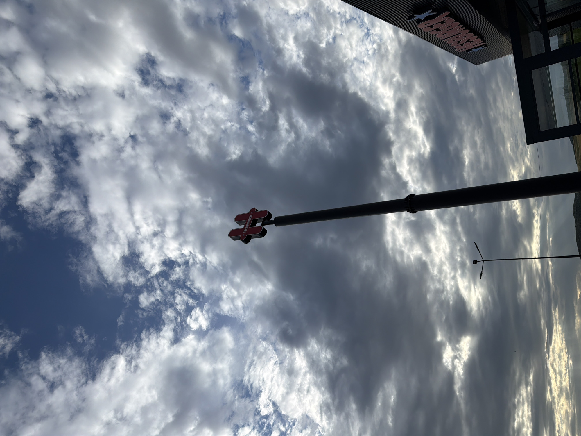 The image depicts a Hesburger restaurant in Polsko Padarevo, Bulgaria, viewed from below against a cloudy sky. The restaurant's sign is partially visible at the top, and a Hesburger logo is seen on a pole. A streetlight is also present. The sky is filled with clouds, illuminated by sunlight from behind.