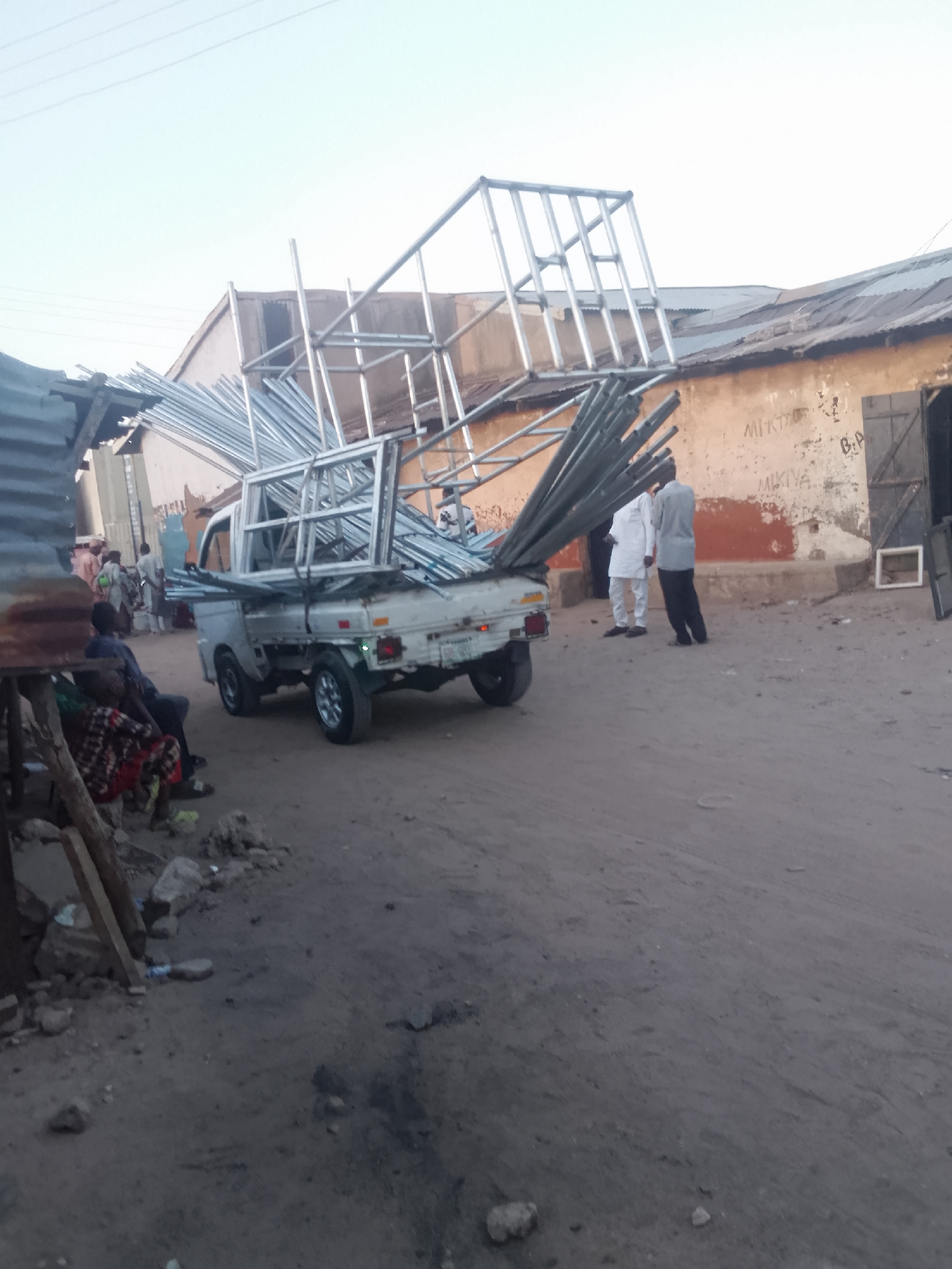 A compact white pickup truck overloaded with aluminum framing and pipes sits on a sandy street in Pantame, Nigeria. Two men stand near the truck, one in white robes and the other in grey. A person is seated near a makeshift wooden structure on the left, partially obscured. The truck is surrounded by dirt and debris. In the background, buildings with aging facades and corrugated metal roofing can be seen. Graffiti is visible on one building. Other people are standing in the distance.
