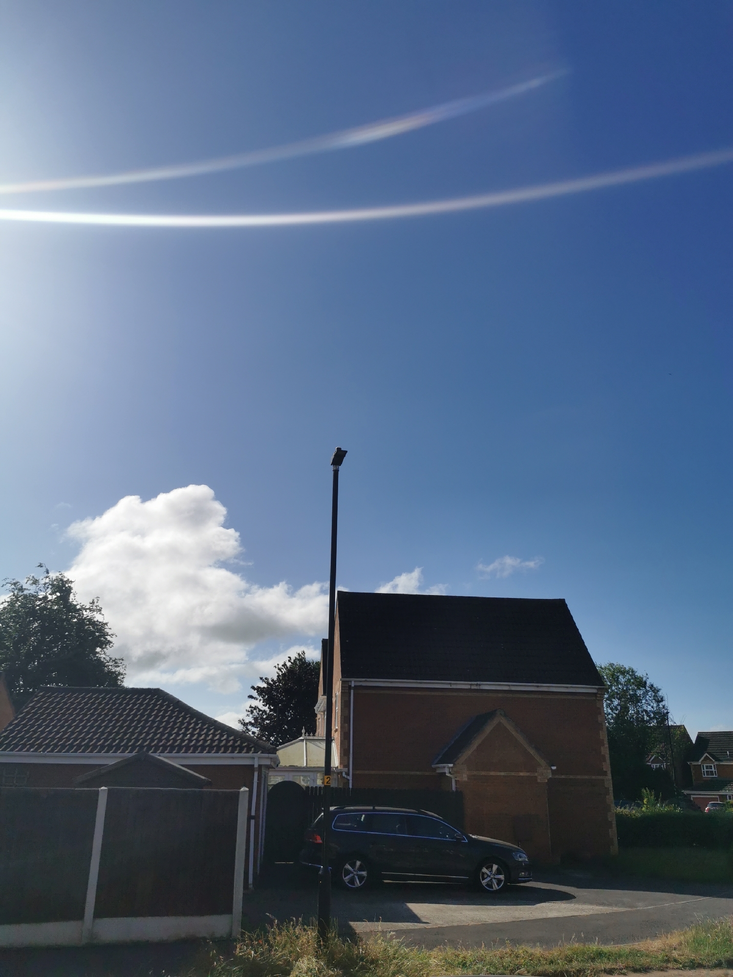 The image shows a suburban street scene in Ripon, United Kingdom, under a clear blue sky with two faint, white contrails visible. A dark-colored estate car is parked on the right side of the frame, in front of a two-story brick house with a dark tiled roof. A tall, dark lamppost stands beside the house. To the left of the house, a smaller, single-story building with a brown tiled roof is partially visible, separated by a dark wooden fence. Trees and other houses are visible in the background. The overall impression is one of a quiet, residential area on a sunny day.