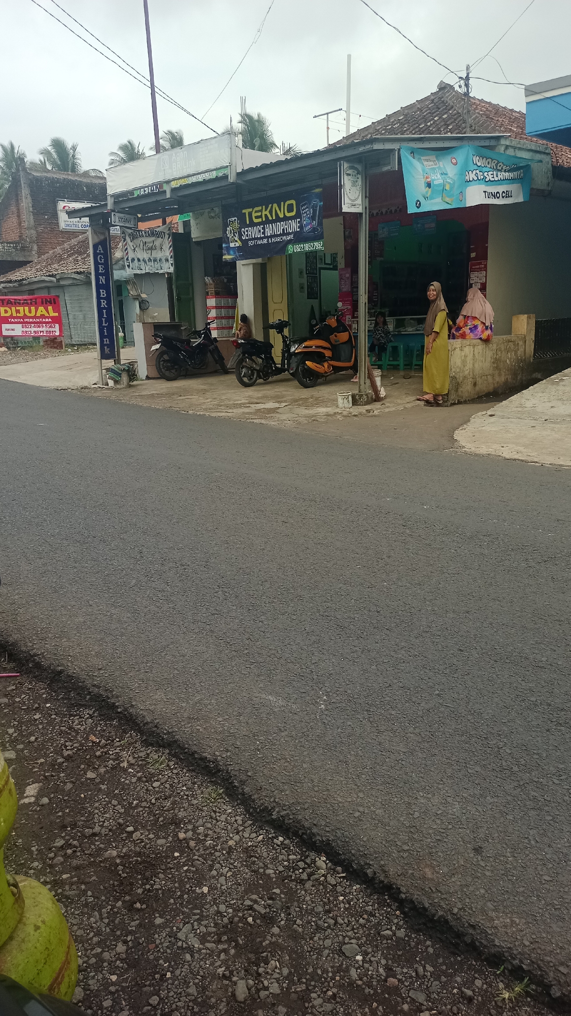 The image depicts a street scene in Karyamandala, Indonesia, featuring a row of shops along a paved road under an overcast sky. The shops are characterized by their corrugated metal roofs and signage, with one prominently displaying "TEKNO SERVICE HANDPHONE SOFTWARE & HARDWARE" and a phone number. Another banner advertises "YOKOR GAK AKTU SELAMATNYA TUKNO CELL". Several motorcycles, including an orange scooter and two black motorcycles, are parked in front of the shops.
Two women are visible. One, wearing a yellow hijab and dress, stands near the shops. Another woman, also in a hijab, sits on a low wall to the right of the shops. The environment suggests a small commercial area in a rural or semi-urban setting, with palm trees visible in the background. The road surface is asphalt, and the area beside the road is a mix of gravel and dirt. A portion of what appears to be gas cylinders is visible in the lower left corner of the image.
