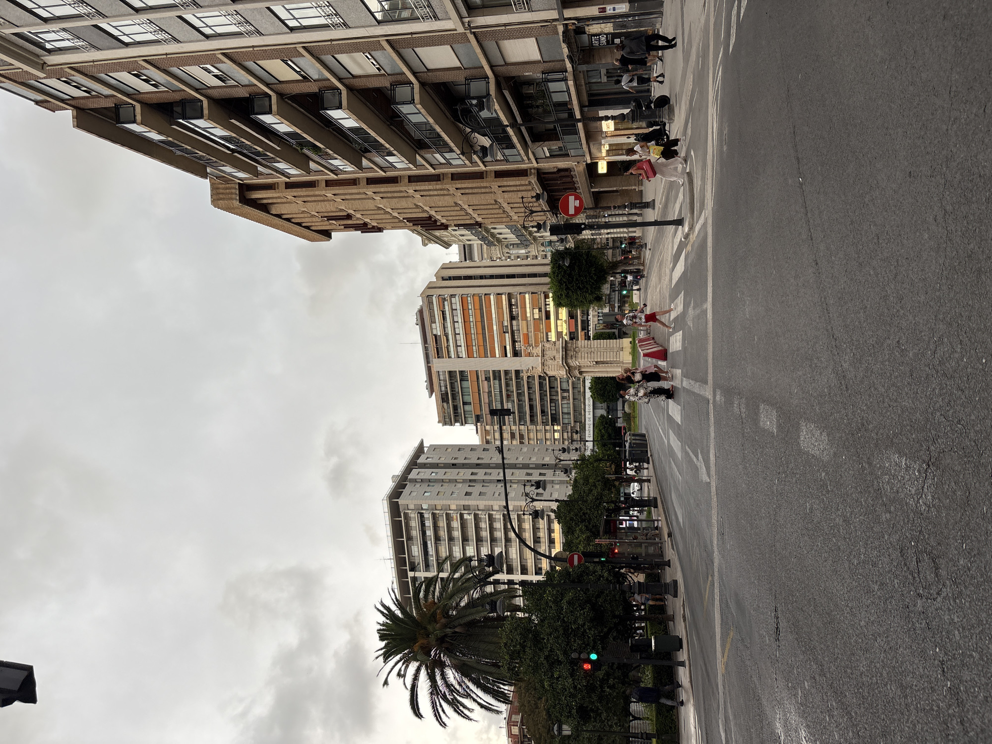 The image depicts a street scene in the city of València, Spain, under an overcast sky. In the foreground, a paved road with pedestrian crosswalks and lane markings dominates the lower half of the frame. Several people are visible crossing the street, including a woman in a white patterned garment, another in a pink dress, and a person in red shorts and a white shirt. To the left of the road, a traffic light shows a green signal, and a palm tree stands tall beside it. Trees and lampposts line the sidewalk. In the background, a series of multi-story buildings with varying architectural styles and colors are arranged along the street. One building features an orange facade with numerous windows. Further back, another tall, light-colored building with many balconies is visible. A grand, ornate architectural structure, possibly a monument or archway, is partially visible in the middle ground to the left. The overall atmosphere is that of a city during daytime, with moderate pedestrian activity.