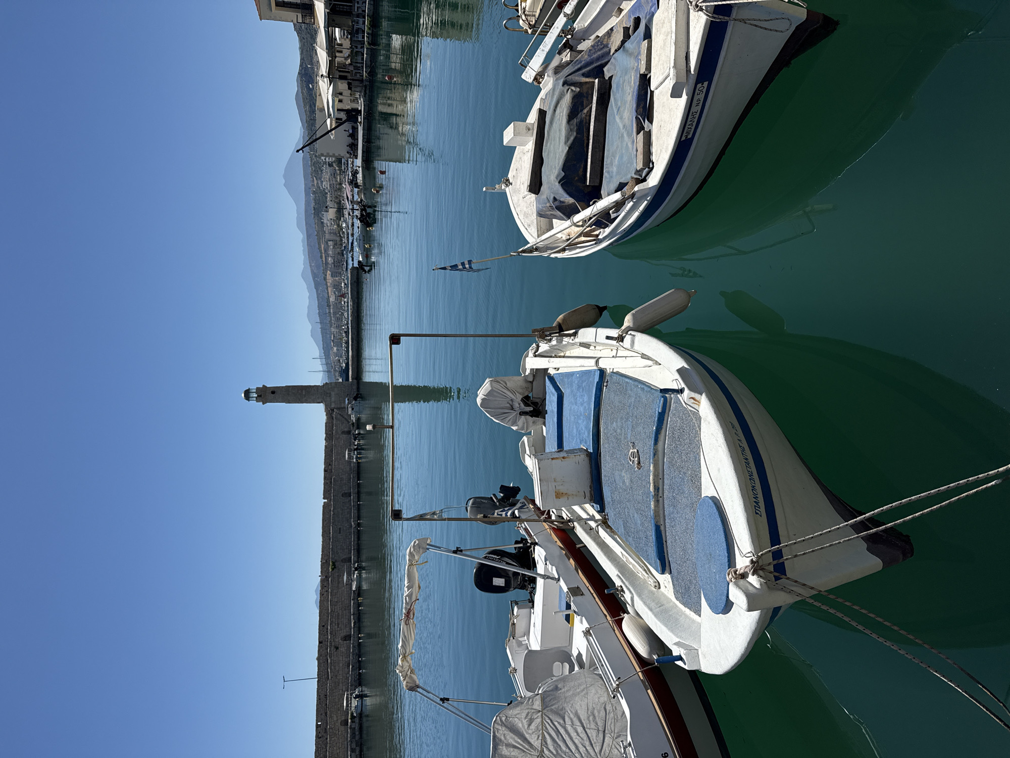 The image shows a view of the Venetian Harbor in Rethymno, Greece. In the foreground, three small boats are docked on the clear, turquoise water. The boat on the left is white with a red stripe along its hull and has an outboard motor. The middle boat is also white, with blue cushions and a canopy. The boat on the right is white with blue trim and has a Greek flag attached to its stern. The middle and right boats are covered with tarps. In the background, a stone breakwater stretches across the harbor, with a lighthouse at its end. Beyond the breakwater, the city of Rethymno can be seen with buildings and mountains in the distance. The sky is clear and blue.