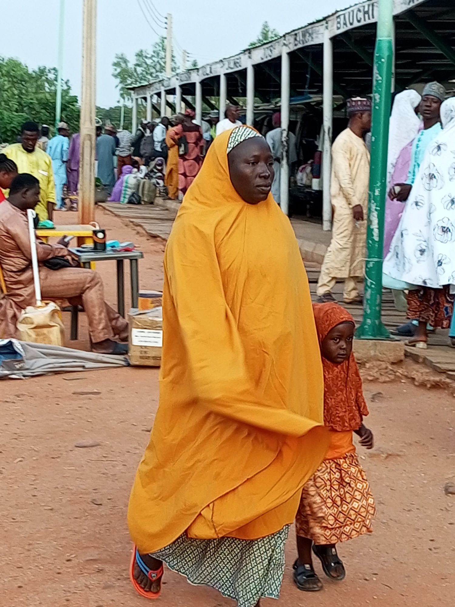 A medium-shot, outdoor, eye-level, daytime photograph shows a bustling scene in the city of Gombe, Nigeria, near a structure labeled "BAUCHI." In the foreground, a woman wearing a large, flowing orange hijab and skirt walks to the left, accompanied by a young child wearing a matching orange headscarf and a patterned skirt, also walking to the left. The woman's face is turned slightly away from the camera, while the child's face is more visible. Behind them, numerous people are visible, some walking, others standing or sitting. The environment appears to be a dusty, open area, possibly a marketplace or transportation hub, with wooden structures and what seem to be goods or luggage in the background. The lighting is diffuse, suggesting an overcast day or late afternoon.