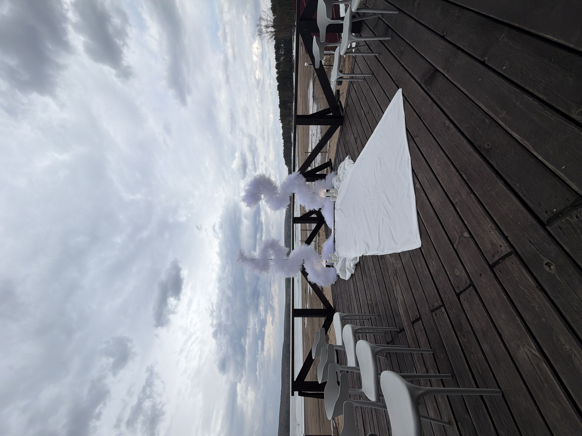 The image displays an outdoor ceremonial setup on a wooden deck overlooking a body of water, likely a lake or wide river, under a heavily overcast sky. The dark brown wooden deck planks run diagonally, dominating the lower and right portions of the frame. Several rows of modern, light gray chairs are arranged on the deck, facing towards the water, suggesting seating for an audience or guests.
In the mid-ground, a white-draped table or platform is positioned. Behind it, closer to the dark wooden railing that separates the deck from the shore, are two prominent white decorative arrangements. These fluffy, cloud-like structures, possibly made of feathers or artificial material, frame a central space, suggesting an archway or focal point for a ceremony. The overall arrangement strongly indicates preparation for an event like a wedding, commitment ceremony, or special celebration.
Beyond the deck and railing, a calm body of water stretches towards a distant landmass, which is covered by a dense treeline, suggesting a forested shore. The sky is entirely covered with thick, gray clouds, indicating an overcast day with subdued lighting. No people are present in the scene. The setting evokes a sense of calm and anticipation, likely a scenic outdoor venue in a location such as Chaikovsky, Russia. There is no visible text in the image.
