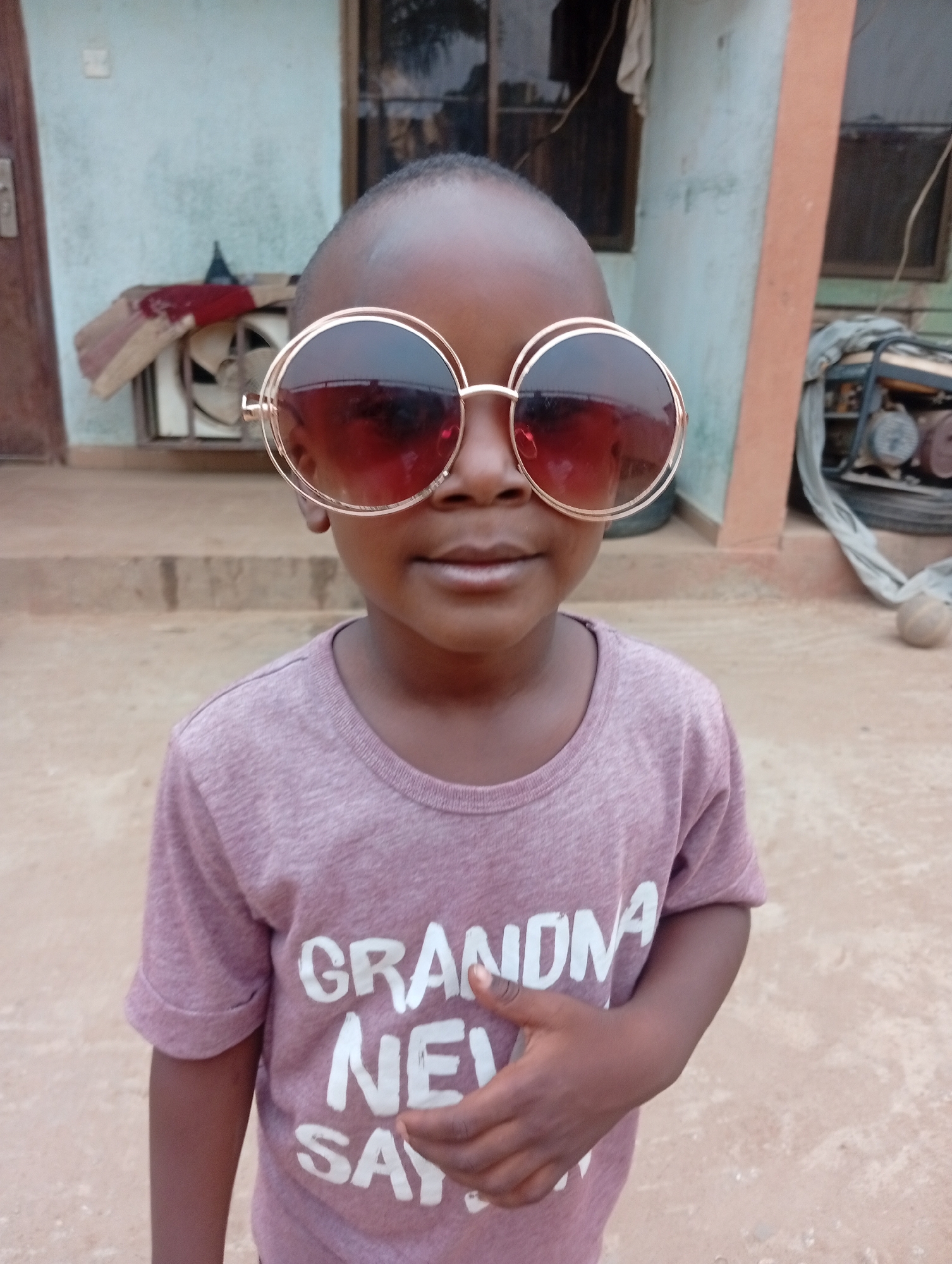 A young child with dark skin is standing in front of a building wearing oversized round sunglasses with a gold frame and dark lenses. The child is wearing a purple t-shirt with the words "GRANDMA NEVER SAY" printed in white letters. The building behind the child has a light blue exterior with an orange column on the right. A closed brown door is visible on the left. Behind the child, there is an electric fan, a generator, and a brown ball on the ground. The setting appears to be a residential area in Sabon Tasha, Nigeria, based on the environmental characteristics.