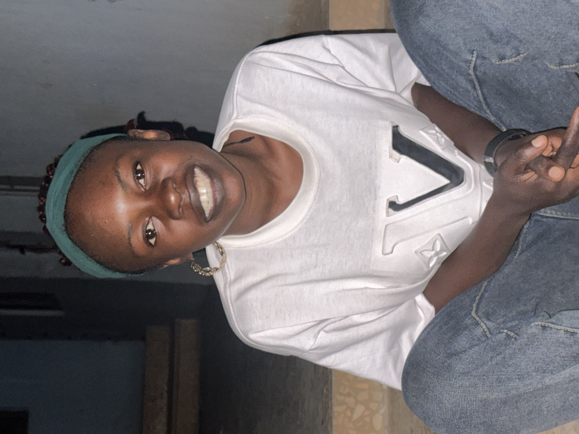 A medium shot captures a young Black woman with dark skin, a green headband, and a white t-shirt with a large "V" logo, sitting on concrete steps. She is smiling, revealing her teeth, and wearing large hoop earrings. Her hands are clasped in her lap, and she wears a black watch on her left wrist. She is dressed in blue jeans. The background is dark and out of focus, with hints of a wall and some stairs. The lighting suggests it may be evening or nighttime. The scene appears to be outdoors, possibly in a residential area or public space within Jos, Nigeria.