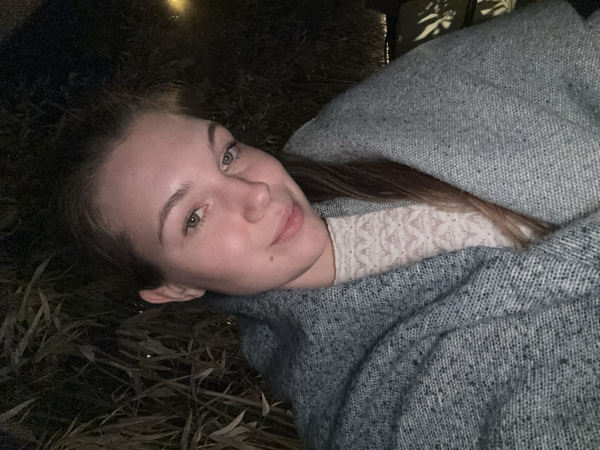 A fair-skinned female is lying on what appears to be dry grass or reeds, covered by a gray speckled blanket. She is wearing a light-colored lace top under the blanket. The environment is dark, illuminated by a single light source in the upper part of the image. The location cannot be determined to be Belgrade, Serbia with the available information.