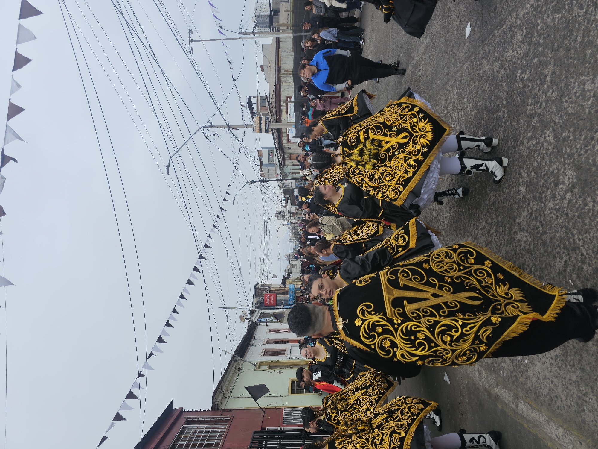 The image captures a lively street scene in Iquique, Chile, likely during a festival or parade. In the foreground, a group of performers, predominantly women, are dressed in elaborate black and gold costumes. These costumes feature ornate embroidered detailing, particularly on capes or ponchos that drape over their shoulders and torsos, with a prominent "V" monogram visible on some. The performers are wearing white knee-high socks and black and white boots. They are walking or dancing in formation down a paved street lined with buildings.
The environment suggests an urban setting. Buildings of varying architectural styles, some appearing older with red siding and barred windows, others more modern, flank the street. Overhead, a complex network of power lines and streetlights crisscrosses the sky, punctuated by strings of triangular flags, alternating between white and dark colors, adding a festive touch to the overcast day. A Coca-Cola advertisement is visible on one of the buildings in the background.
On the sidewalks and edges of the street, a crowd of spectators watches the procession. They are dressed in casual, everyday clothing. The overall atmosphere appears to be one of community celebration and cultural expression. The sky is a muted gray, indicating cloudy weather.