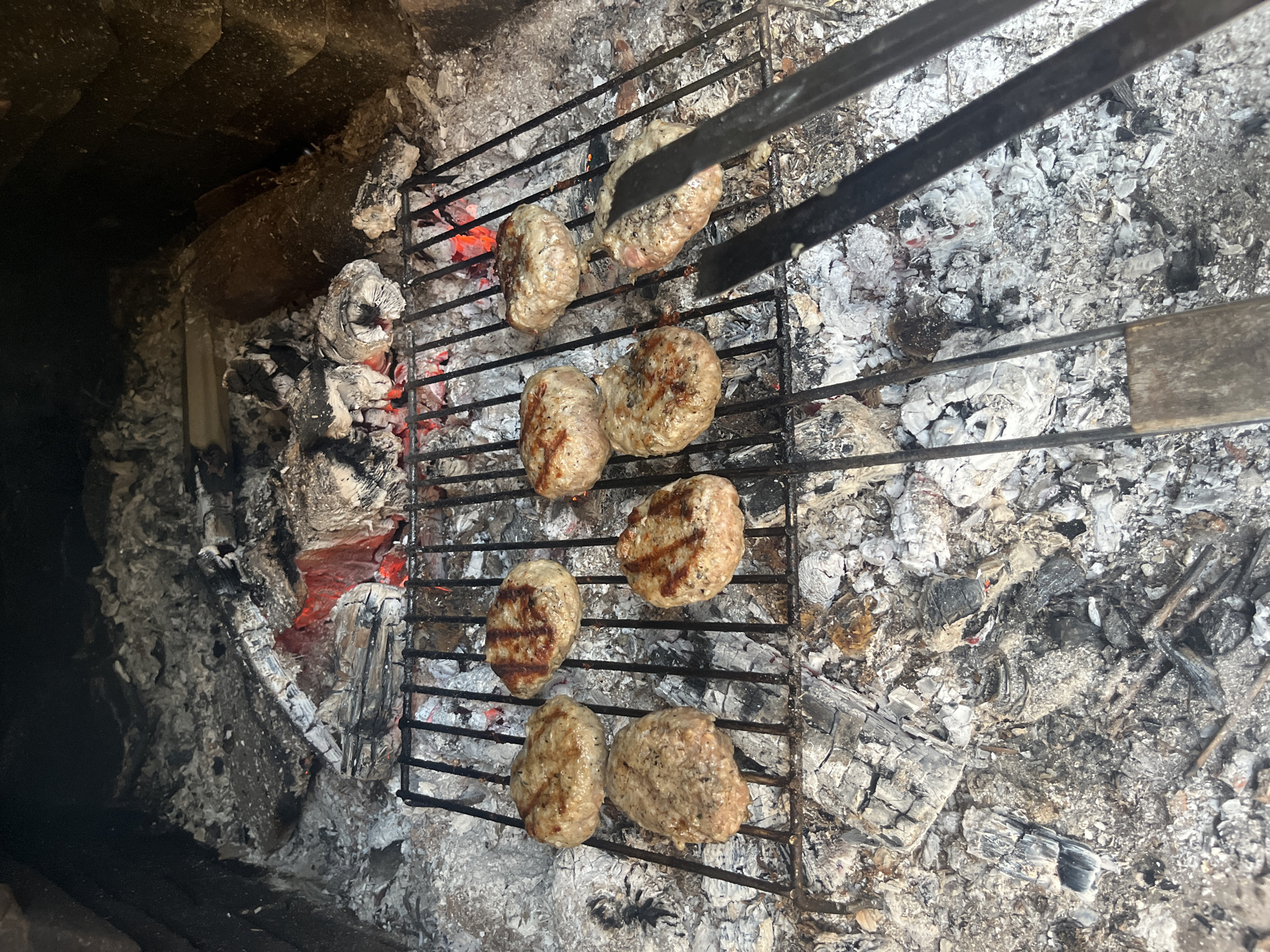The image shows a grill with several small, oval-shaped meat patties cooking over a bed of hot coals and ash. A pair of metal tongs is visible, presumably used for turning or moving the patties. The cooking setup appears to be inside a brick structure, perhaps a small oven or fireplace. The location is Vladaya, Bulgaria.