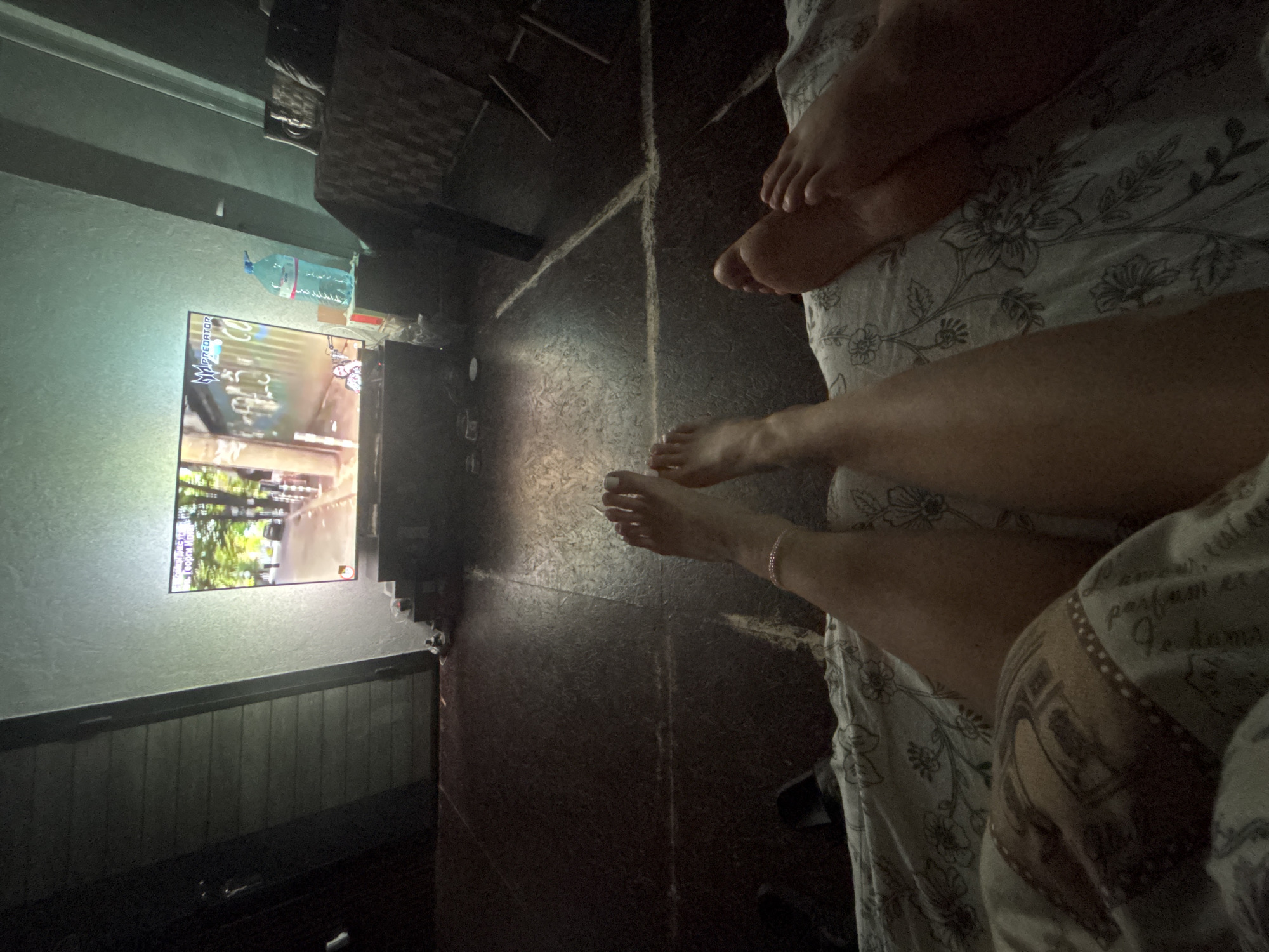 An indoor scene shows two people in bed watching a television. The viewer's perspective is from the bed, focused on the lower bodies and feet of the two individuals. The bedding has a floral print. A television screen is visible on the wall across the room, displaying content with green and white hues. To the side of the TV is a plastic water bottle. The floor between the bed and the television has a textured, dark surface. The location is Belopoptsi, Bulgaria.