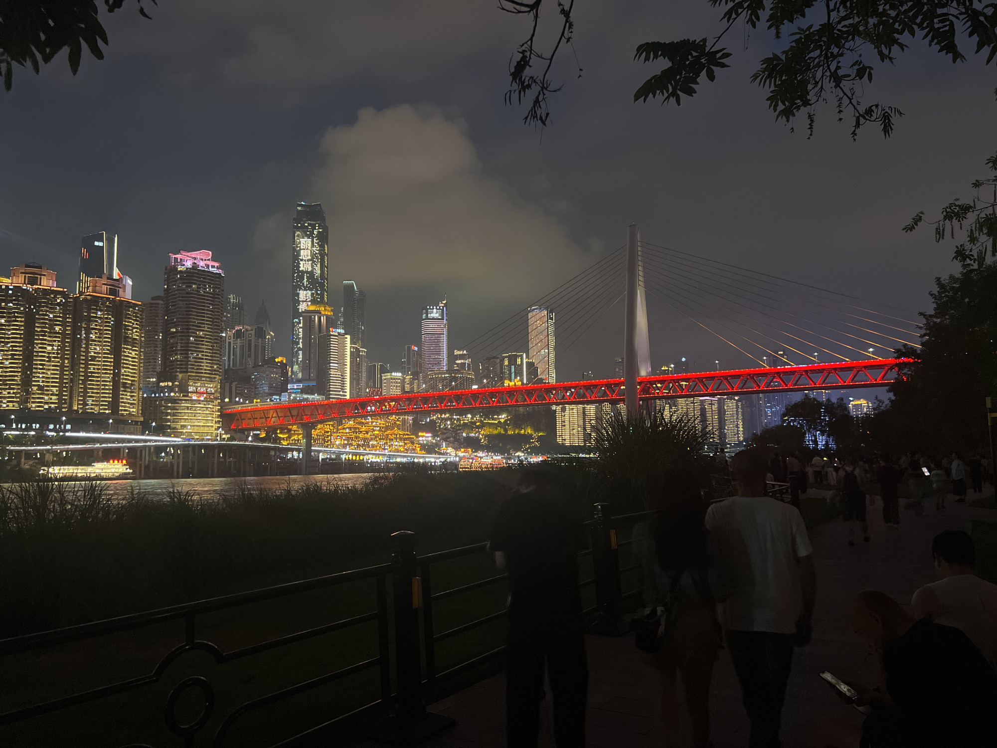 The image depicts a nighttime scene in Yuzhong District, China, showcasing a brightly lit cityscape and a prominent red bridge spanning a body of water. Tall, illuminated skyscrapers dominate the background, their windows glowing with warm light. The bridge, a striking feature, is adorned with red lights that create a vibrant glow across its structure and reflect on the water below. In the foreground, silhouetted figures of people are visible, some walking along a pathway bordered by a dark fence, while others are seated. The overall atmosphere is one of urban nighttime activity, with the city lights creating a lively and dynamic backdrop.