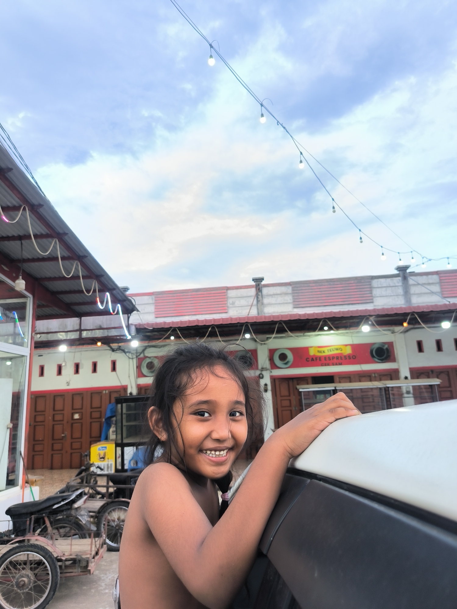 This outdoor photograph, taken in Sukamulia, Indonesia, features a young girl as the central subject. She is positioned in the foreground, looking directly at the camera with a wide, infectious smile that reveals her teeth. Her dark hair appears wet or damp, and her shoulders are bare. She is leaning on the white roof and black trim of a vehicle, which partially fills the lower right section of the frame.
The background depicts a semi-urban scene with a series of buildings, possibly shops or houses, under a cloudy sky. These structures feature red-tiled roofs, white walls, and dark brown doors with small, square windows. Some buildings are adorned with decorative, colorful strip lights. A prominent red sign on one building reads "REX TEUWO," "CAFE ESPRESSO," and "CERIAM." Overhead, strings of illuminated bare light bulbs stretch across the frame, suggesting the time of day is late afternoon or early evening. To the left, a motorcycle with a sidecar or modified cargo attachment is visible. The overall mood is cheerful, accentuated by the girl's happy expression amidst a lively local setting.