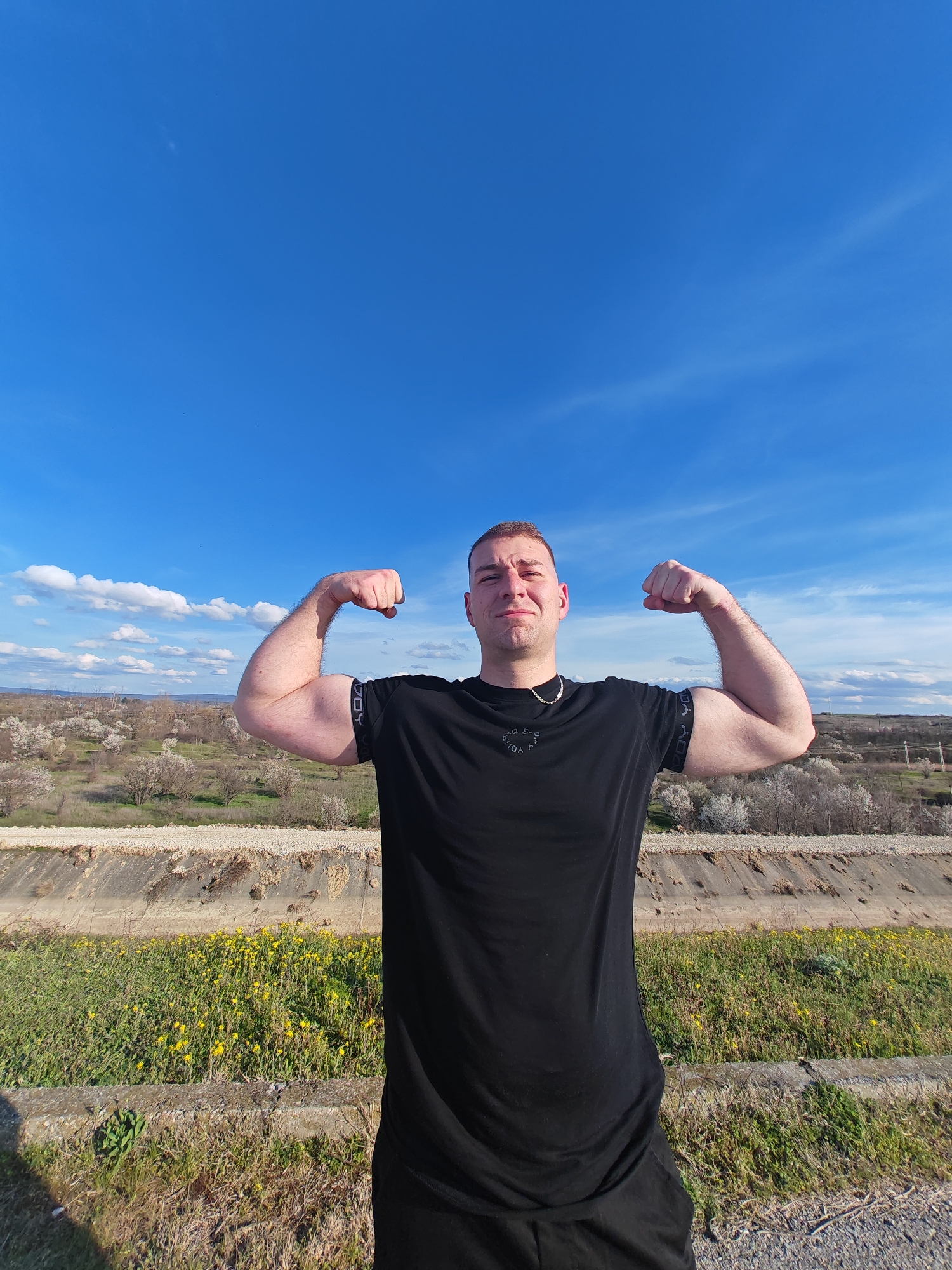 A man poses outdoors with a blue sky as the backdrop. He is wearing a black t-shirt and black pants. He is flexing both arms, revealing defined biceps. He has short hair and a gold chain around his neck. The background features a landscape with green grass, yellow flowers, a concrete structure resembling a channel or wall, and flowering trees in the distance. The scene is likely located in a rural area outside of a city. Based on the information, the location is near Shumen, Bulgaria.
