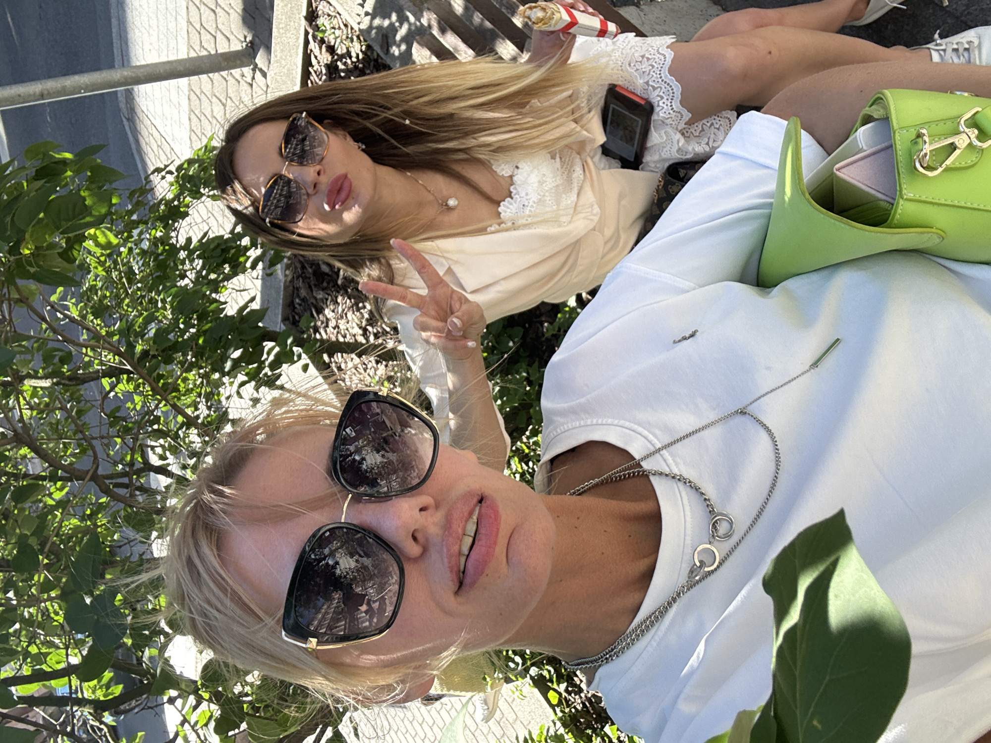 Two women are pictured outdoors on a sunny day, likely in Saint Petersburg, Russia, seated on a bench near a tree. The woman in the foreground, on the left, is wearing a white t-shirt, large black sunglasses, and silver necklaces. She is looking directly at the camera with a slight smile. The woman in the background, on the right, is wearing a light-colored dress with lace detailing and large sunglasses. She is making a peace sign with her hand and sticking her tongue out playfully at the camera. She is holding a food item in her other hand and has a phone on her lap. The environment appears to be a park or street side area with paved ground and vegetation.