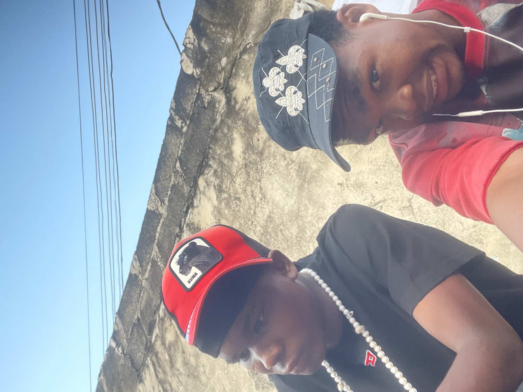 In Ado, Nigeria, two young men are pictured outdoors under a clear blue sky. The man on the left wears a red baseball cap with a black panther logo and a black t-shirt, adorned with a pearl necklace. He is looking away from the camera. The man on the right sports a black baseball cap with embroidered white fleur-de-lis symbols and a red t-shirt, wearing earbuds. He is looking directly at the camera with a grimace. Behind them, a textured, weathered concrete wall runs horizontally, with several utility wires stretched across the sky above it. The ground appears to be a light-colored, rough surface.