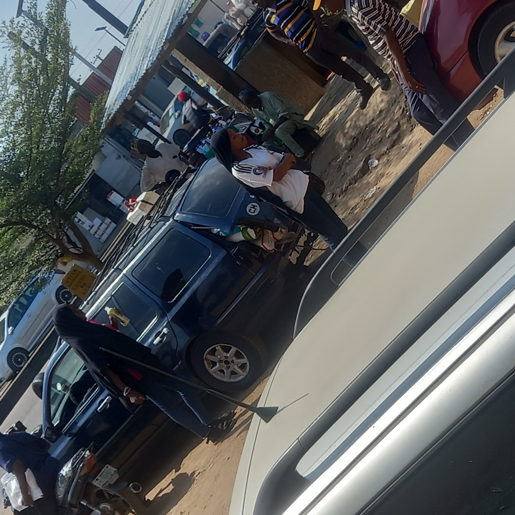 The image shows a street scene in Sardauna, Nigeria. There are several people present, including a woman in a white shirt and dark pants leaning against a dark-colored van. Another woman in a black head covering and jeans is holding an object resembling a mop or squeegee near the front of the vehicle. Several other people are standing and sitting nearby. The environment includes various vehicles, buildings with corrugated metal roofs, and trees. Some items are stacked on the roof of the van. The overall impression is a typical street scene with a mix of vehicles, pedestrians, and vendors.
