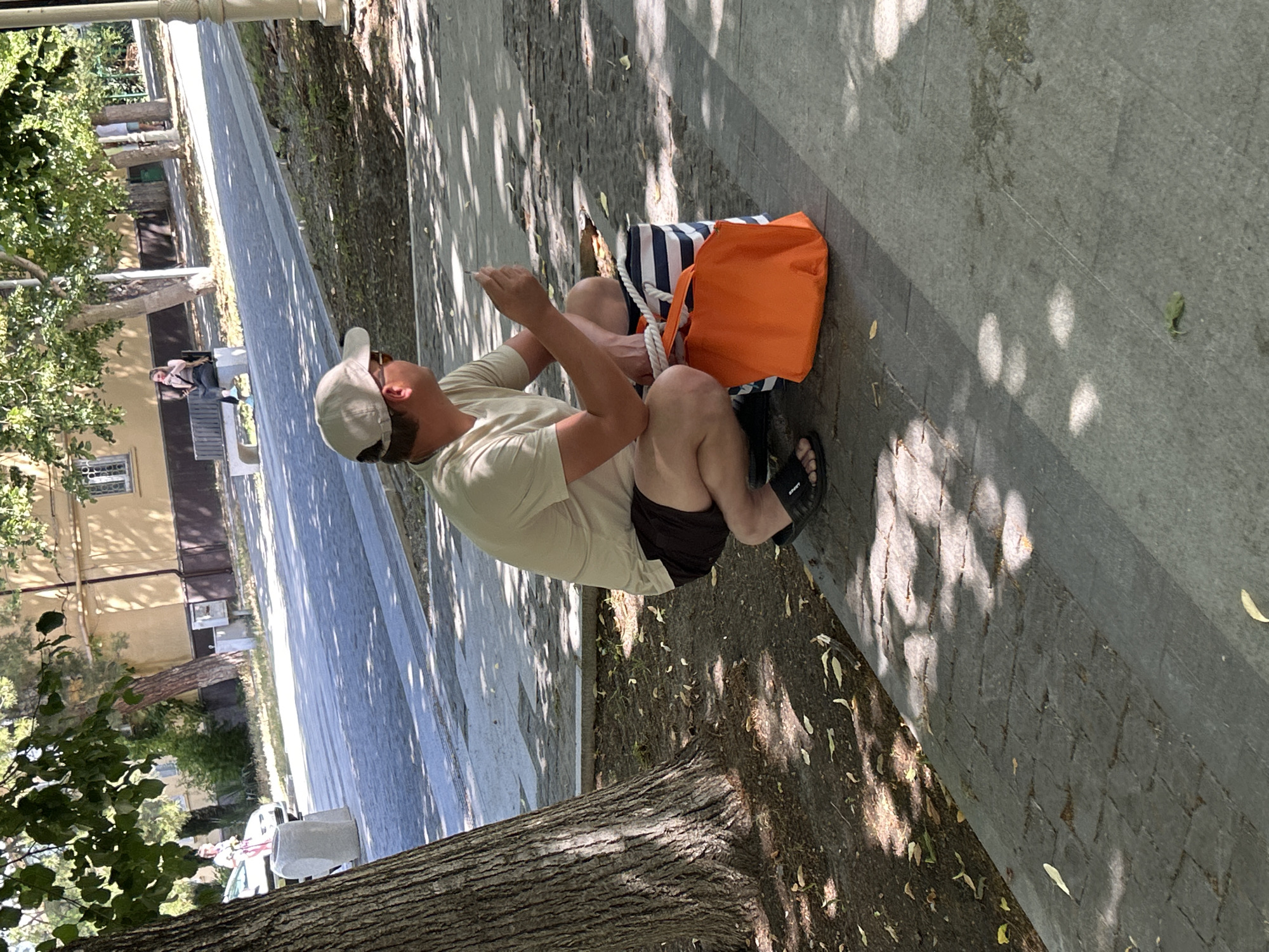 In Gelendzhik, Russia, a man wearing a light-colored t-shirt, dark shorts, a beige baseball cap, and black sandals is squatting on a cobblestone sidewalk. He is holding a phone in his right hand and looking to the right. Beside him are two bags: an orange tote bag and a blue and white striped bag with a white rope handle. The man is positioned near the base of a large tree with dappled sunlight filtering through its leaves onto the ground. In the background, a building with yellow siding and brown trim is visible, along with a woman sitting on a park bench. The environment appears to be a park or public walkway with trees and paved areas.