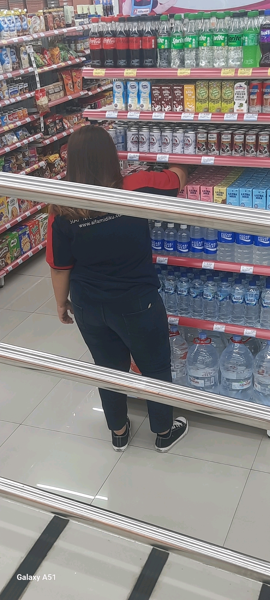 A person wearing a blue shirt with red accents and dark jeans is standing in a supermarket aisle in Wlingi, Indonesia. They are facing away from the camera, looking towards shelves stocked with various beverages. The shelves display bottled water, soft drinks like Coca-Cola and Sprite, and boxed milk. In the foreground, a silver metal railing runs horizontally across the image, partially obscuring the view of the person and the shelves. The floor is tiled. The overall scene depicts a retail environment with products displayed for sale.