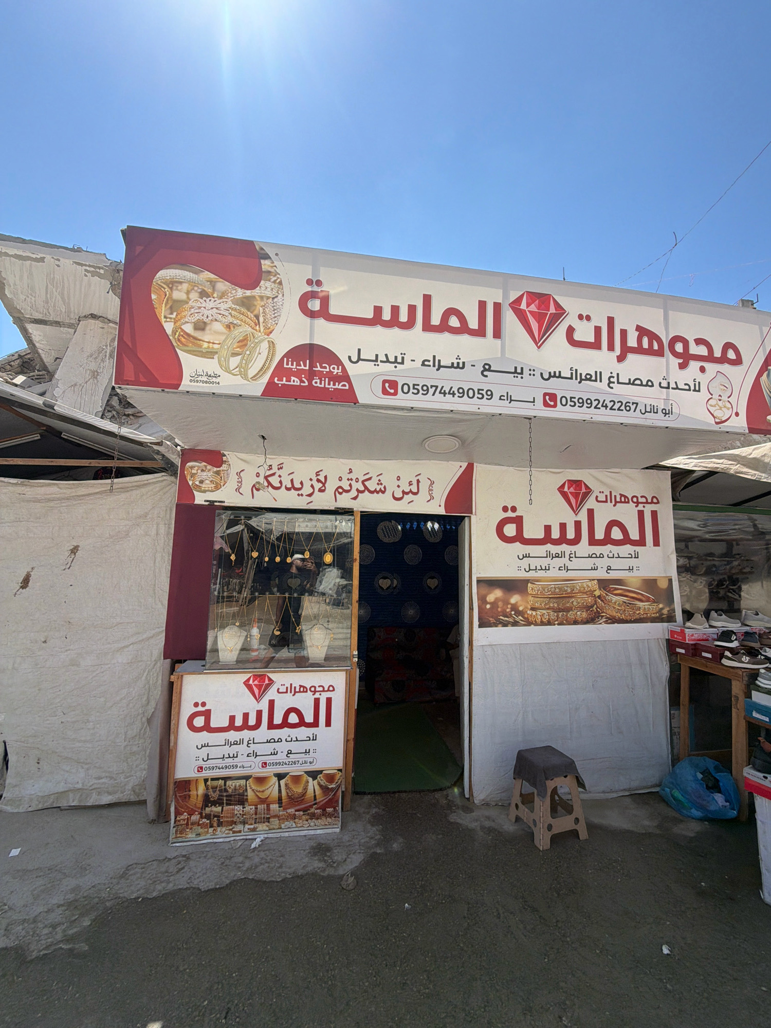 The image shows the exterior of a jewelry store in the city of Gaza, Palestinian Territories. The store has a large sign with the name "الماسة" (Al-Masa) and a diamond logo, indicating it sells jewelry. Text on the sign includes "لأحدث مصاغ العرائس" (for the latest bridal jewelry), and services like buying, selling, and repairing gold. Two phone numbers are also visible. The store appears to be located in a damaged area, with the remnants of a collapsed building visible in the background. The storefront features a display window with various gold necklaces and rings. A small wooden stool with a dark fabric cover is placed outside the store, along with a blue plastic bag and some shoe boxes. The ground is unpaved and appears wet in some areas. The sky is clear and blue with bright sunlight.