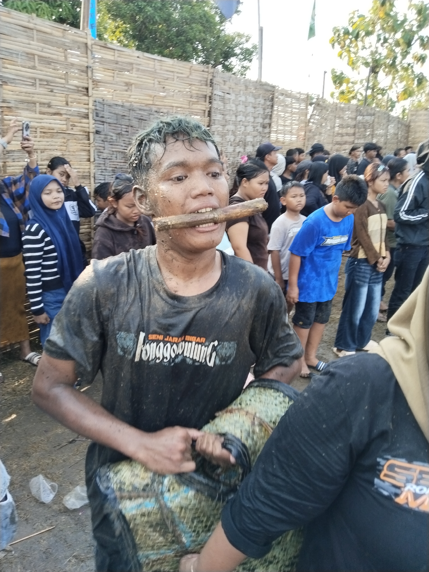 The image shows a man with a wooden stick through his mouth. He is wearing a dirty t-shirt and holding what appears to be a woven basket. The background shows a crowd of people, a bamboo fence, trees, and flags. The image was taken in Kedawung, Indonesia.