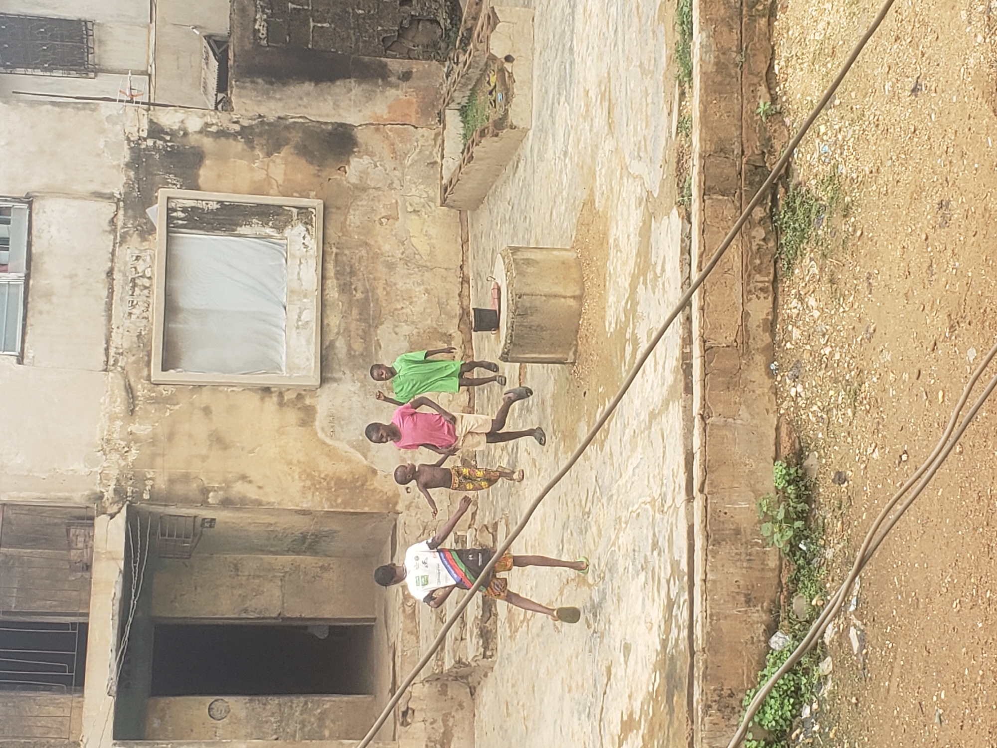 The image depicts a courtyard in Alalubosa, Nigeria, adjacent to a weathered building with deteriorated facade. Four children are present. One wears a white shirt and patterned shorts, one a pink shirt and khaki shorts, another a green shirt and black shorts, and the last is shirtless with patterned shorts. The children appear to be playing, possibly running or skipping. A concrete well is centrally located in the courtyard, with a container inside. Utility wires traverse the upper right of the frame. The courtyard is paved with cracked concrete. Adjacent to the pavement is a dirt area with sparse vegetation.