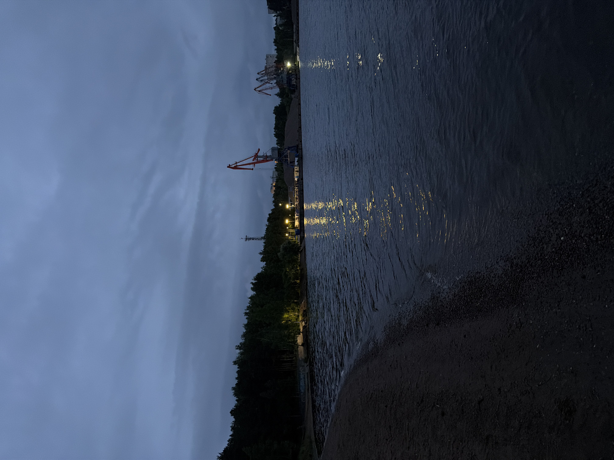 The image depicts a nighttime scene of a waterfront in Chaikovsky, Russia. The sky is overcast with dark blue-grey clouds, occupying the upper two-thirds of the frame. In the foreground, a dark, sandy beach with a narrow strip of pebbles curves along the left edge, meeting a large body of dark water that fills the lower right quadrant. Ripples are visible on the water's surface, reflecting numerous elongated streaks of artificial yellow light emanating from the far bank.
Along the distant bank, a dense line of dark green trees runs horizontally across the midground. Several bright, yellow streetlights illuminate portions of the shoreline behind these trees. Above the treeline on the left, a tall, slender communication tower or mast is visible. Further right, a busy port area is evident, featuring at least two large gantry cranes with prominent red booms and white/grey support structures. One crane, positioned more centrally, is taller and more distinctly lit. Behind the cranes, large piles of granular material, likely aggregates, are stacked. Further to the right, other cranes and industrial structures are visible, with the faint outlines of distant city buildings discernible on the horizon behind the port. No people are visible in the scene.