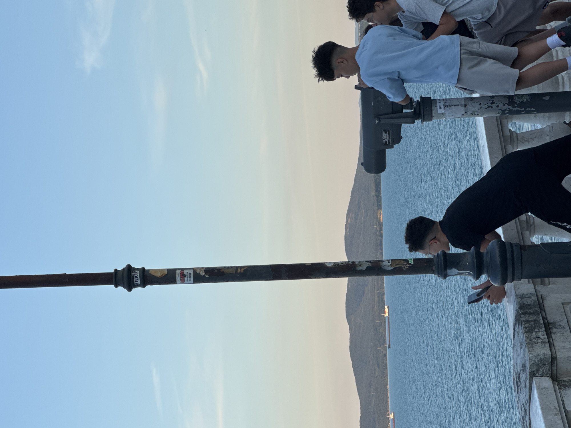 A group of young people are standing on a seawall overlooking the sea. In the foreground, a tall, dark lamp post with stickers on it stands to the left of the frame. A young person in a black t-shirt is holding a phone, looking out at the water. To the right, a young person in a light blue t-shirt and grey shorts is looking through a large coin-operated telescope. Another young person in a grey t-shirt is standing next to them, also looking out at the sea. In the distance, a chain of low, green hills forms the coastline, and several cargo ships are visible on the water. The sky is a clear blue with some faint clouds. The setting is likely a promenade or viewpoint in Burgas, Bulgaria, given the coastal landscape and presence of public viewing telescopes.