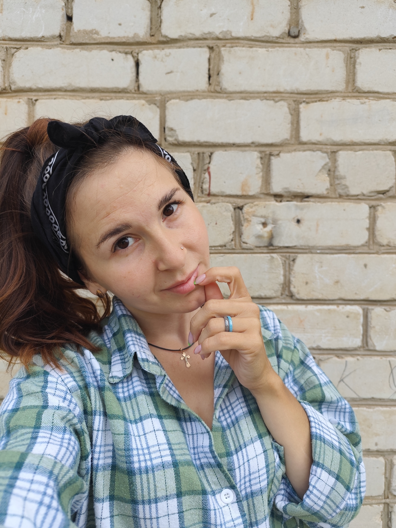 A young woman with brown hair tied back with a black bandana poses in front of a white brick wall. She is wearing a green and white plaid shirt and a gold cross necklace. Her right hand is raised to her face, and she is wearing two rings on her right index finger. The lighting is natural and appears to be daylight. The background is a plain, light-colored brick wall, which shows signs of weathering and some graffiti. The image seems to be taken outdoors. The location is not determinable from the visual information provided.