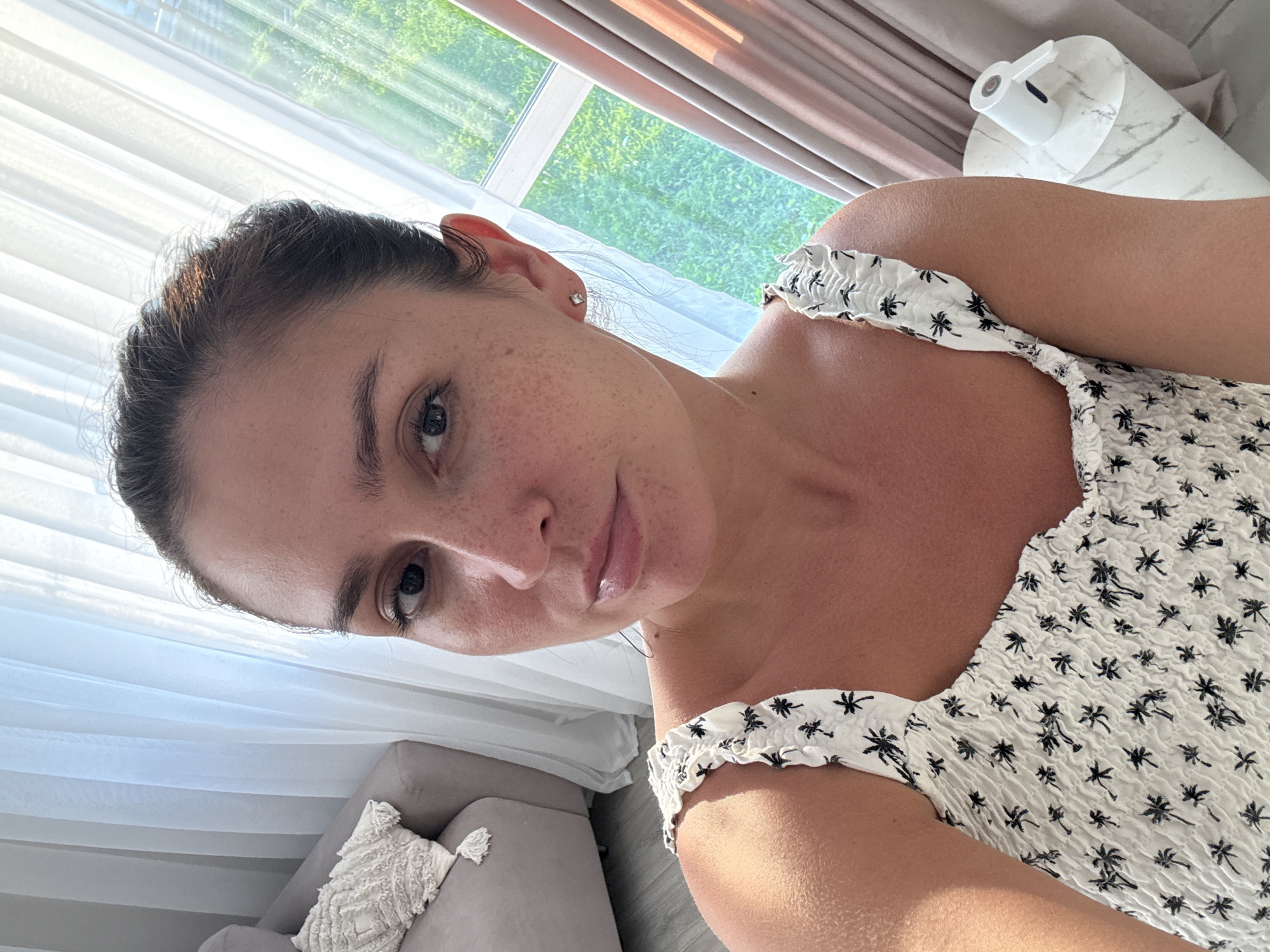 A woman is taking a selfie in a brightly lit room. She has brown hair pulled back, freckles on her face, and is wearing a white dress with a palm tree print. In the background, sheer white curtains are drawn across a window, and a grey sofa with a textured pillow is visible. To the right, a modern side table with a white cylindrical object sits next to light pink curtains, and green foliage can be seen outside the window.