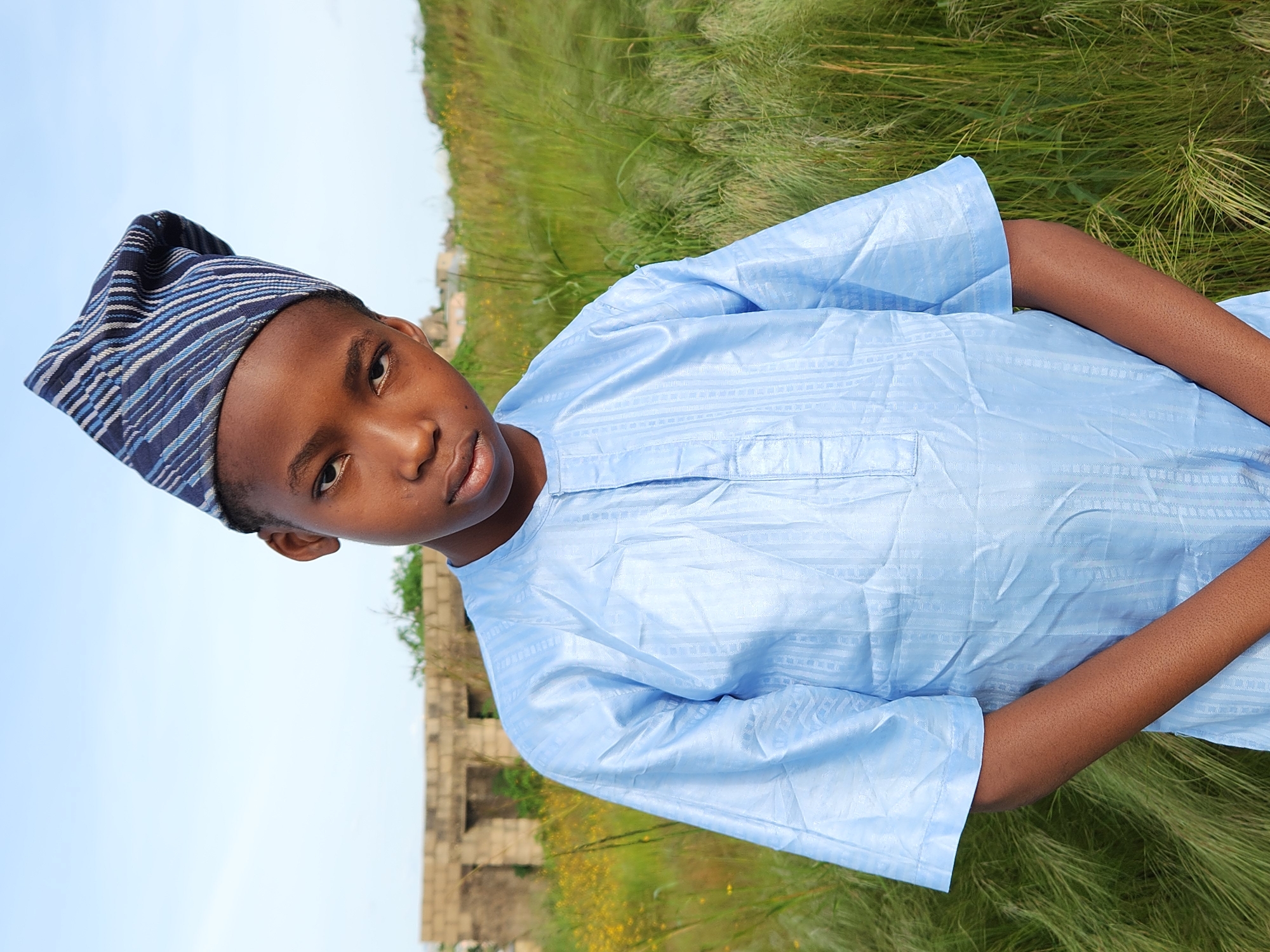 A young boy wearing a light blue traditional outfit and a striped Kufi hat is pictured outdoors. He is looking towards the camera with a neutral expression. The background consists of tall green grass and a clear blue sky. In the distance, partially obscured by grass, are the ruins of a stone structure. The lighting suggests it is daytime, likely overcast or late afternoon, with soft shadows. The overall scene appears to be in a rural or semi-rural setting, with the ruins hinting at a historical or abandoned site. The grass is lush and tall, indicating a potentially temperate season or region. The boy's attire suggests a cultural context, possibly within Jos, Nigeria, where such clothing is common.