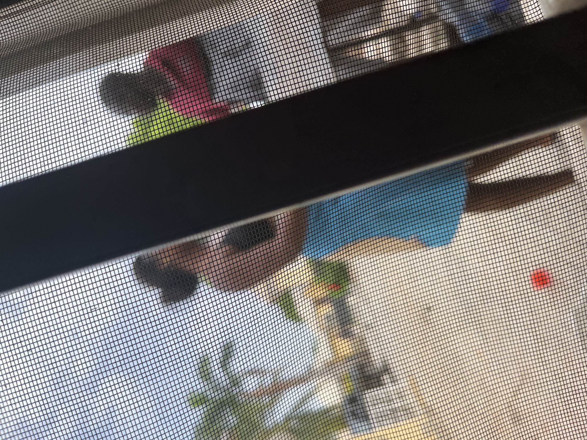 A view through a screen mesh depicts a scene involving people and possibly objects outdoors. One individual appears to be wearing a blue garment and another a pink one. A red circular object is visible in the lower right corner of the image. Vegetation is also discernible. It is not possible to determine the location or provide specific details about actions or interactions from this image.