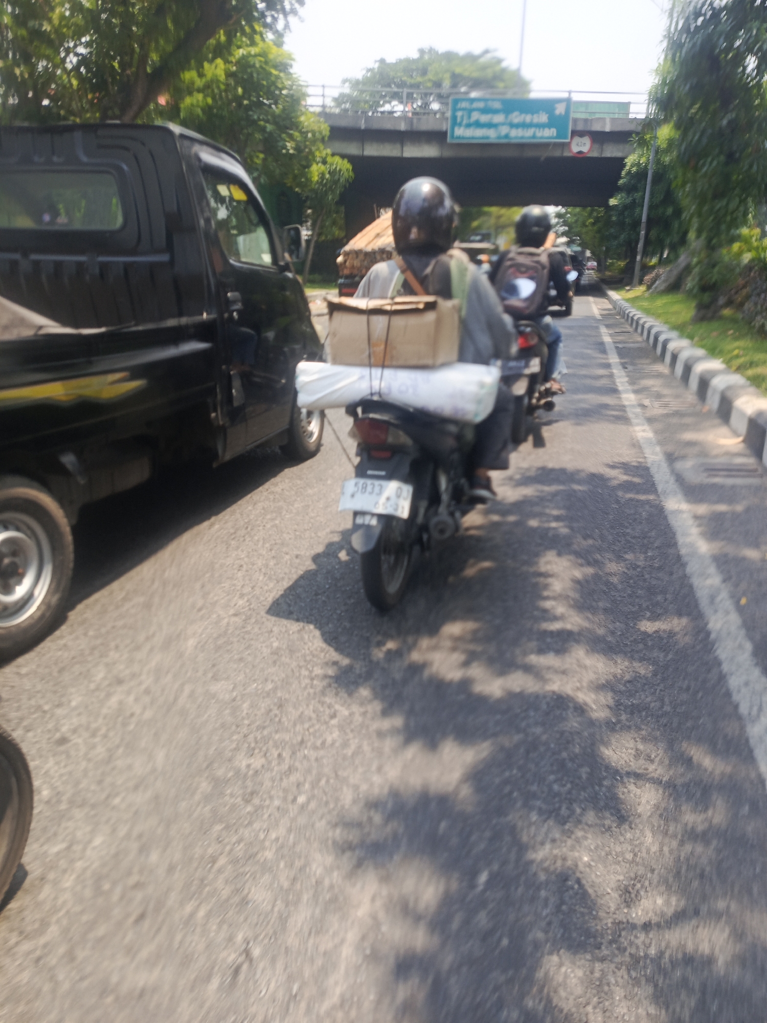 The image depicts a street scene in Surabaya, Indonesia. A black pickup truck is parked on the left side of the road. To the right of the truck, two individuals are riding motorcycles. The first motorcycle, closer to the truck, has a person wearing a black helmet and a backpack. The second motorcycle, further ahead, has two people, both wearing helmets, with the person in the back wearing a backpack. The motorcycles are carrying packages. In the background, there is an elevated road or bridge with traffic signs indicating directions to "Tj. Perak/Gresik" and "Malang/Pasuruan". The road is paved, and there is a grassy area with trees along the right side. The lighting suggests it is daytime, with shadows cast on the road.