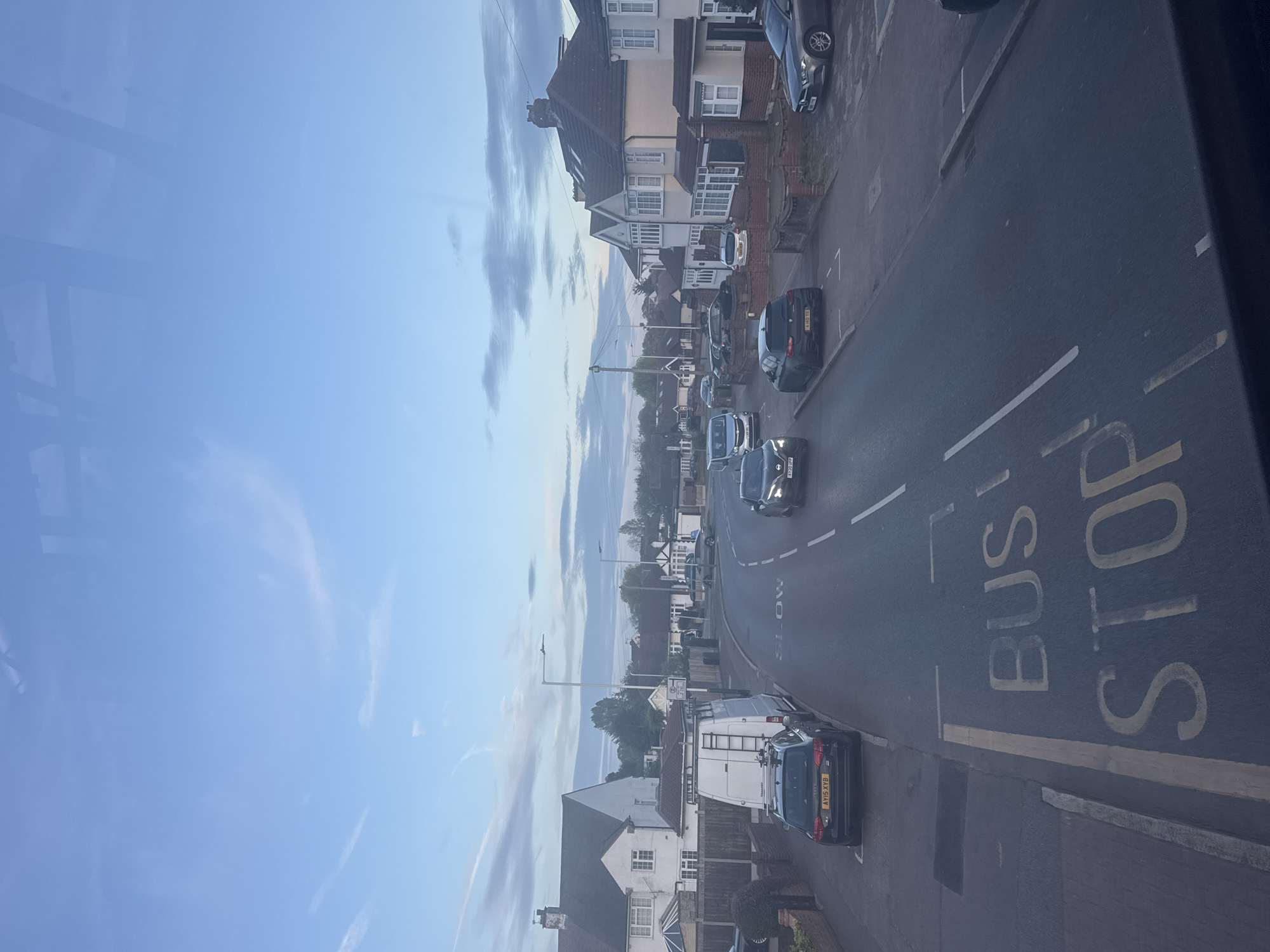 The image captures a residential street scene in London, United Kingdom, likely during daytime under an overcast but bright sky with scattered, wispy clouds. The perspective is from an elevated position, possibly from a window, looking down the street. Reflections, possibly from a window pane or frame, are visible in the upper left corner of the sky.
On the right side of the road, a row of two-story terraced or semi-detached houses with brick and rendered facades and pitched roofs line the street. Several cars are parked along both sides of the road, and more cars are visible driving or parked further down the street. Notable vehicles include a black van with a ladder rack (license plate AH15 XYB) near the foreground, a black car (LJ60 YXF), a silver car (H46 YXA), and another dark car (AV67 YXD) in the distance. The presence of UK license plates and typical British housing architecture confirms the location context.
The asphalt road surface features clear white and yellow road markings. Prominently displayed in the foreground are the large yellow painted words "BUS STOP," with a thick yellow line extending beneath them, indicating a designated bus stopping area. Further down the road, the word "SLOW" is partially visible, also painted in white on the road. Yellow lines run along the curbs, indicating parking restrictions. A tall, slender streetlight pole is visible in the middle distance, and trees and bushes add greenery to the scene, especially between houses and further down the road.
There are no people or specific activities taking place, presenting a calm and ordinary view of a suburban street. The lighting suggests it is either morning or late afternoon, as the light is soft, but it is not dark.