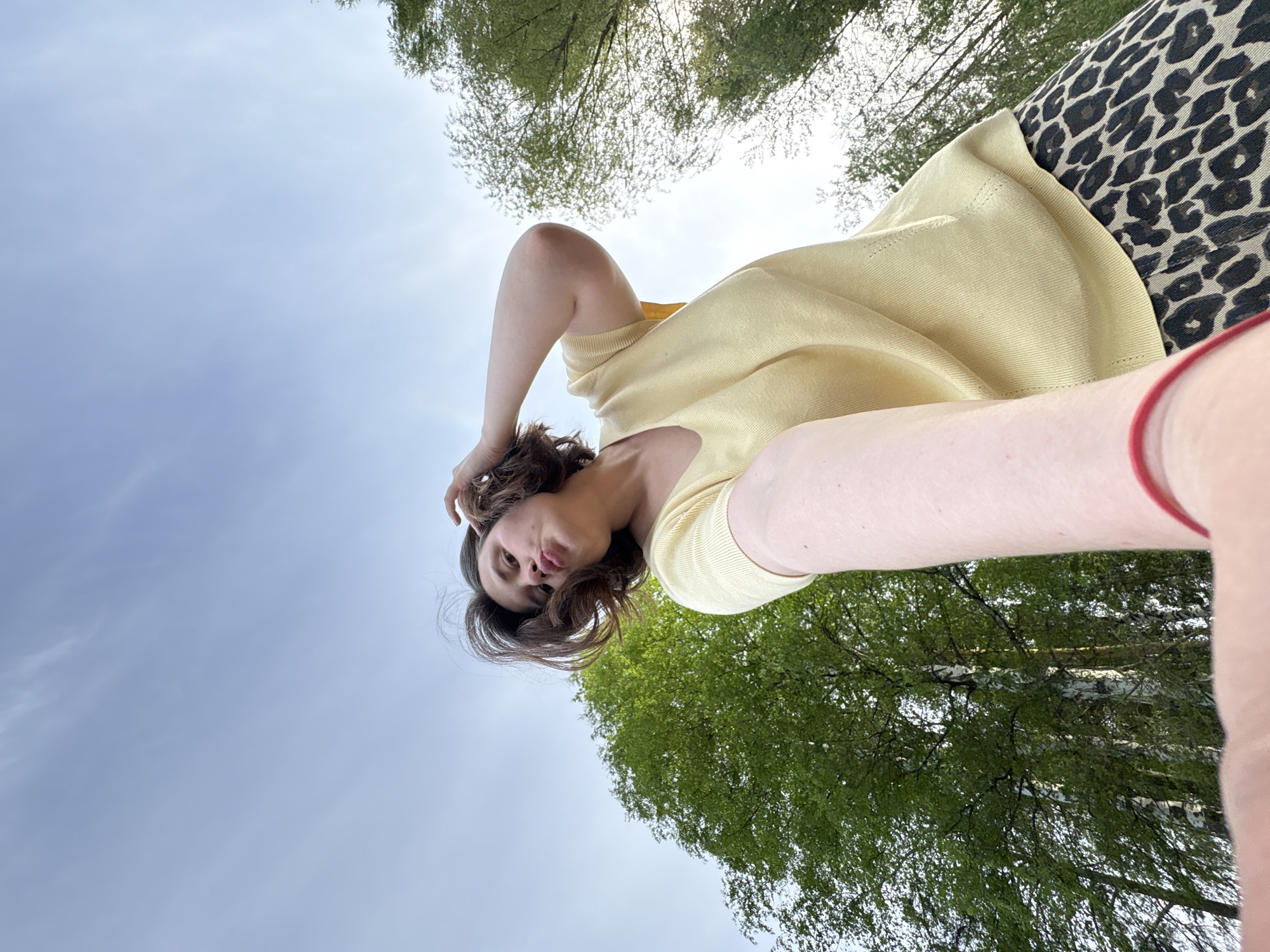 The image is an upward-angled selfie of a woman standing outdoors. She has brown curly hair and wears a light yellow sweater and leopard-print pants. Her left hand is raised to her hair. The background includes trees with green leaves and a light blue sky with some cloud cover.
Given the request, it is impossible to confirm the exact location of the photo, but the prompt states that it should be within the city of Chaikovsky, Russia.
